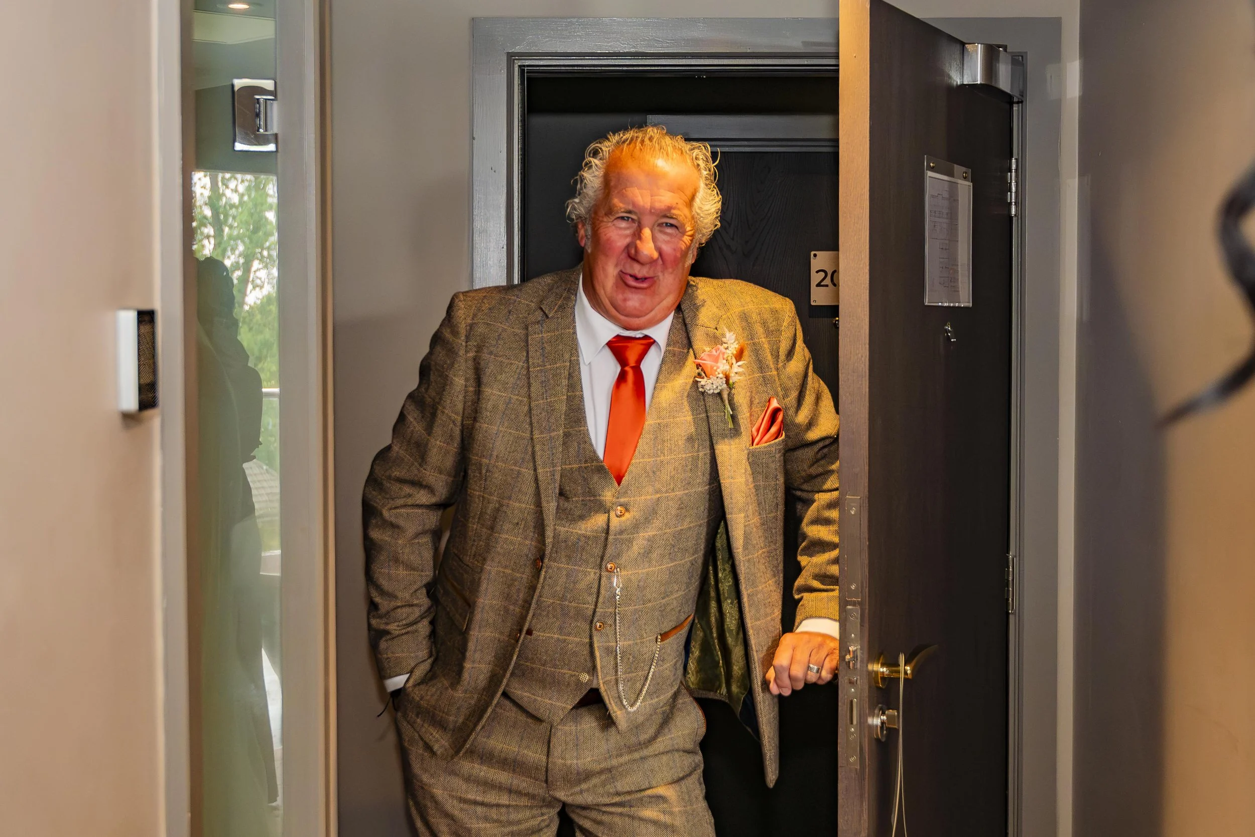 Man with gray hair dressed in a brown plaid suit, white shirt, and orange tie standing at the door of a room, smiling and looking at the camera, with a boutonniere on his lapel.
