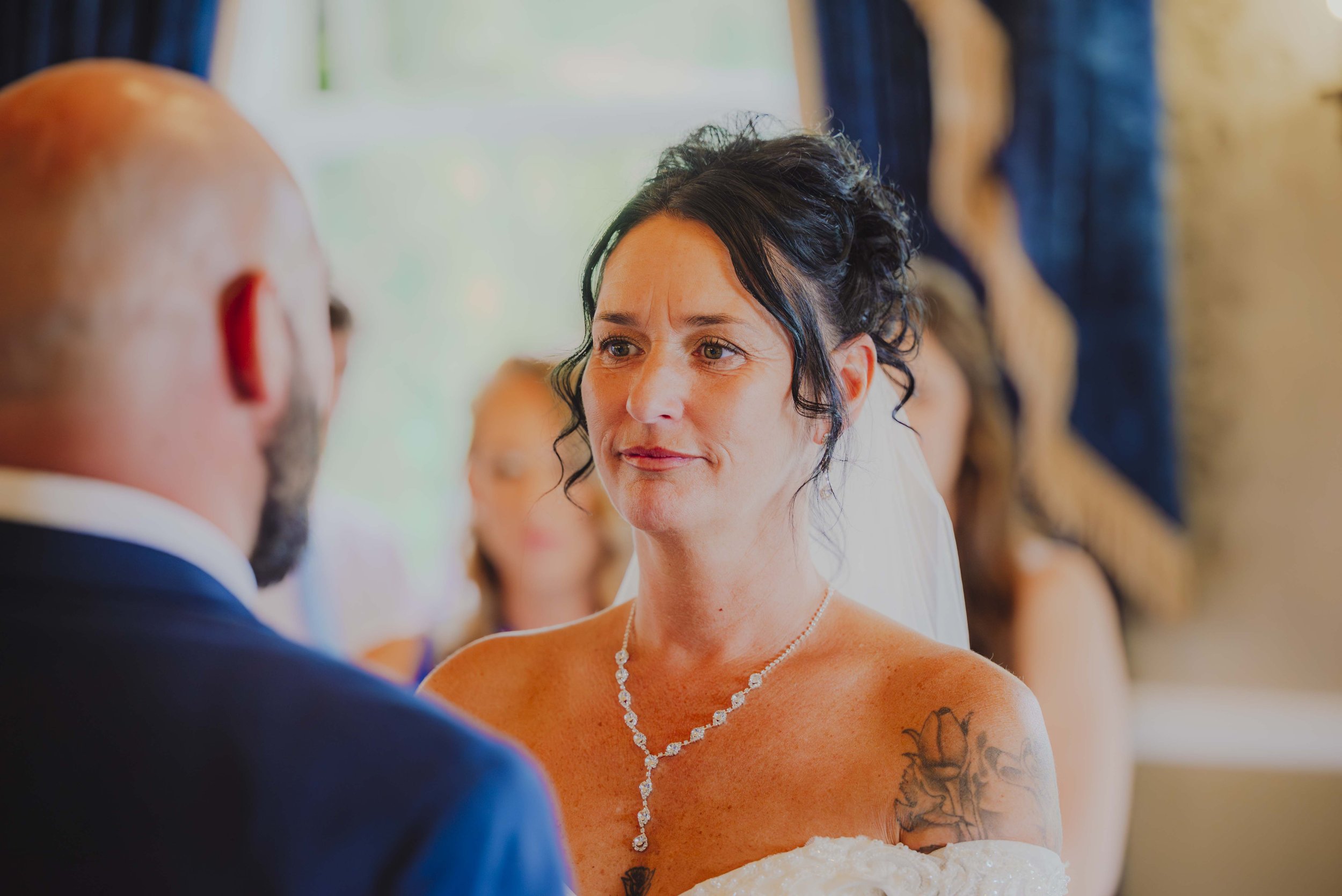 A woman with black curly hair, wedding dress, and tattoo on her shoulder, looking at a man during a wedding ceremony.
