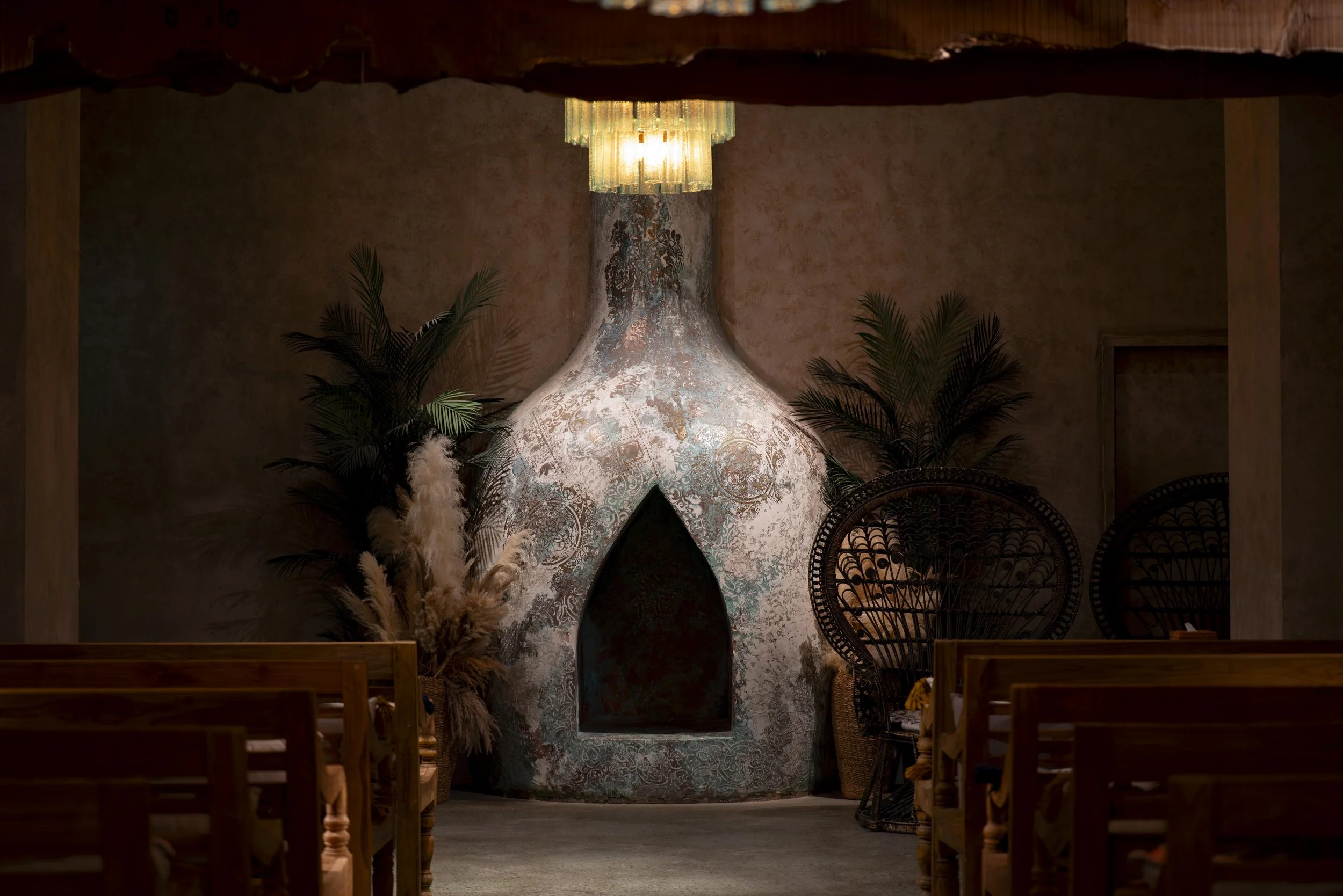 Interior of a room with a large, textured chimney-like structure at the center, decorated with intricate patterns. A chandelier hangs above, and plants and decorative chairs surround the area.