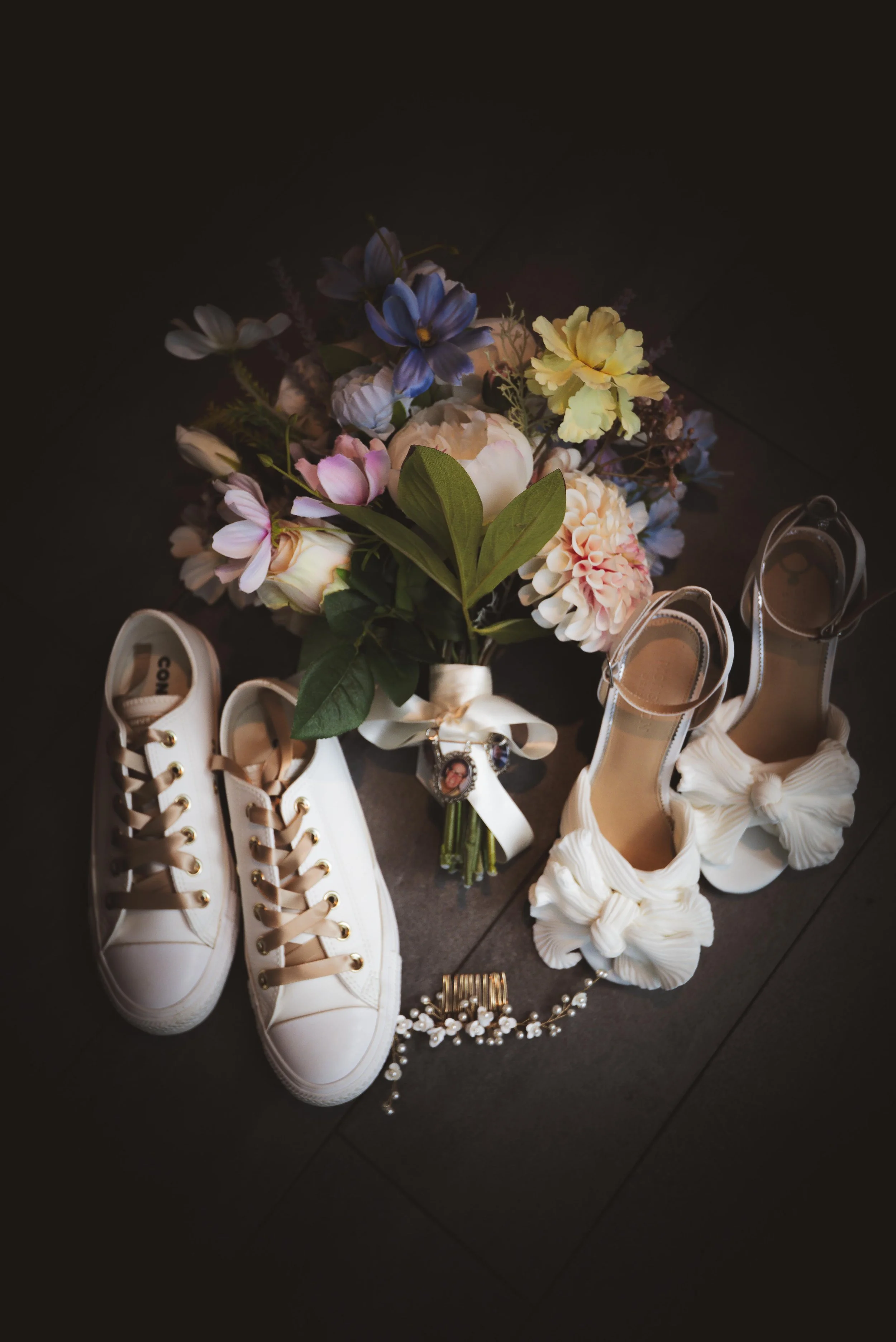 A bouquet of colorful flowers, a pair of white sneakers with beige laces, white high heels with large bows, and a pearl bracelet are arranged on a dark floor.