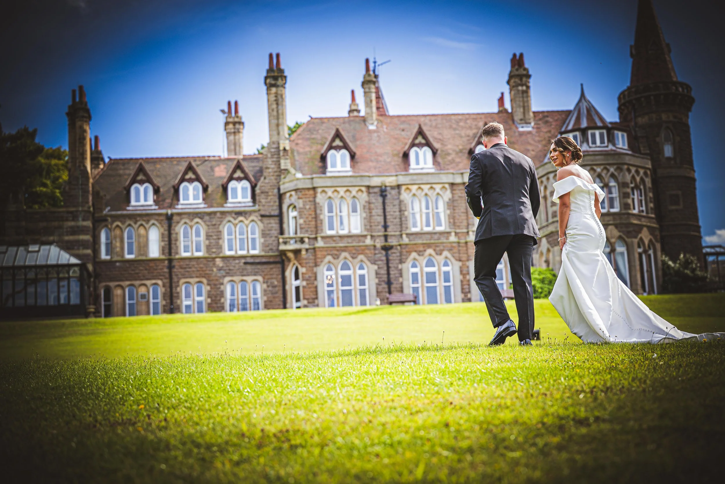 A bride and groom walk on a grassy lawn in front of a large, historic stone mansion during their wedding day.