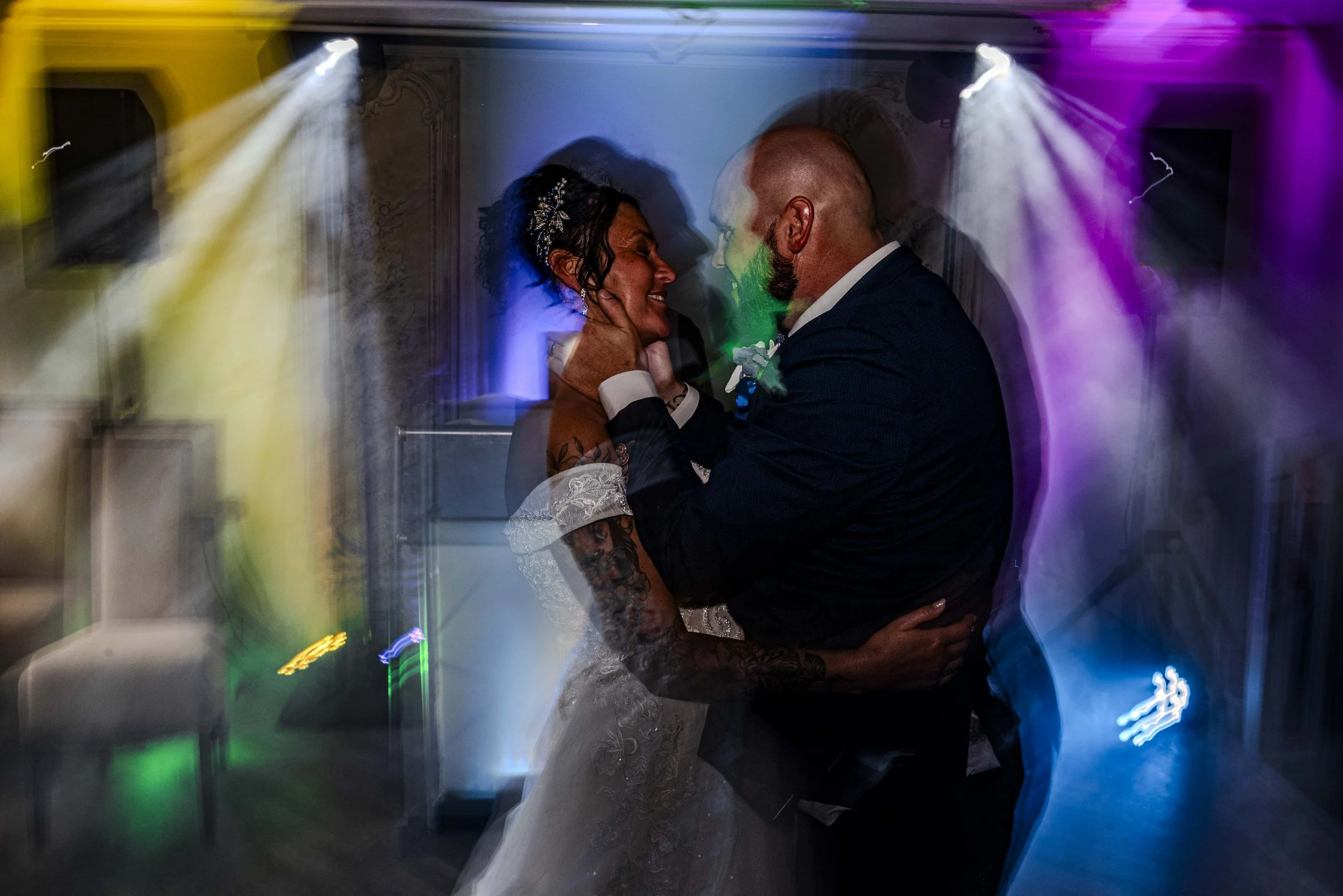 A bride and groom dancing close together at their wedding reception, surrounded by colorful lights and a lively atmosphere.