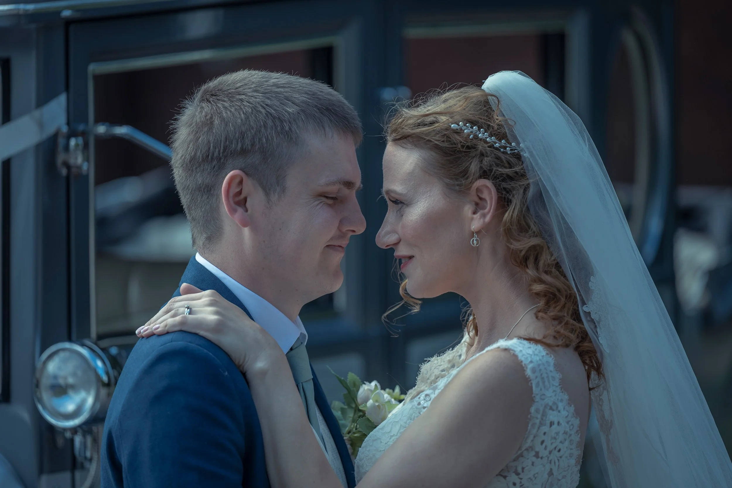 A bride and groom are standing close together, smiling and looking into each other's eyes, with the bride holding the groom's face and the groom gently holding her waist, in front of a vintage car.