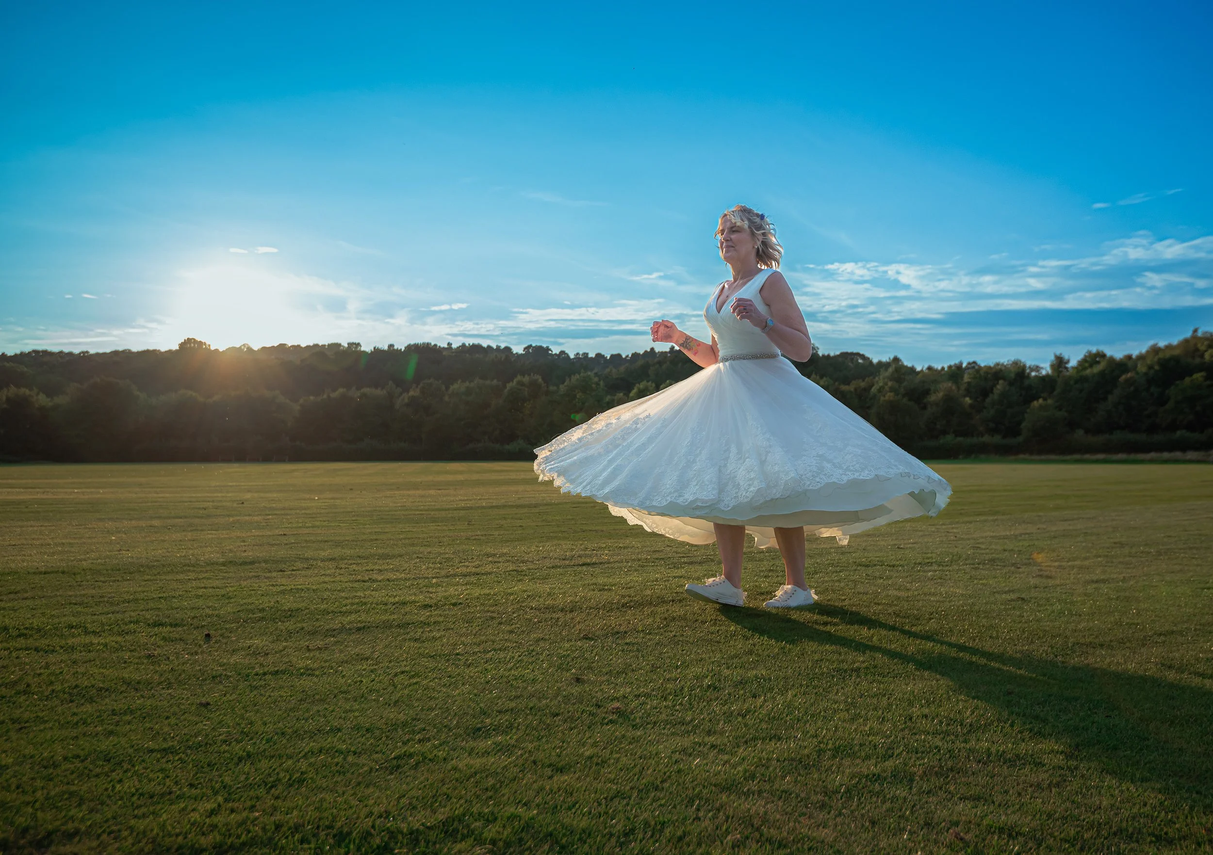 A woman in a white wedding dress and sneakers is spinning in a grassy field during sunset, with trees and a blue sky in the background.