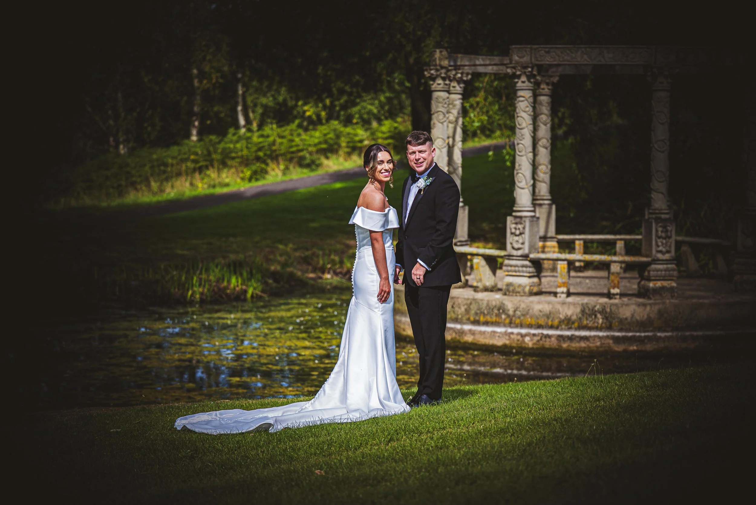 A bride and groom standing side by side outdoors near a pond with a stone gazebo in the background, dressed in wedding attire, smiling.