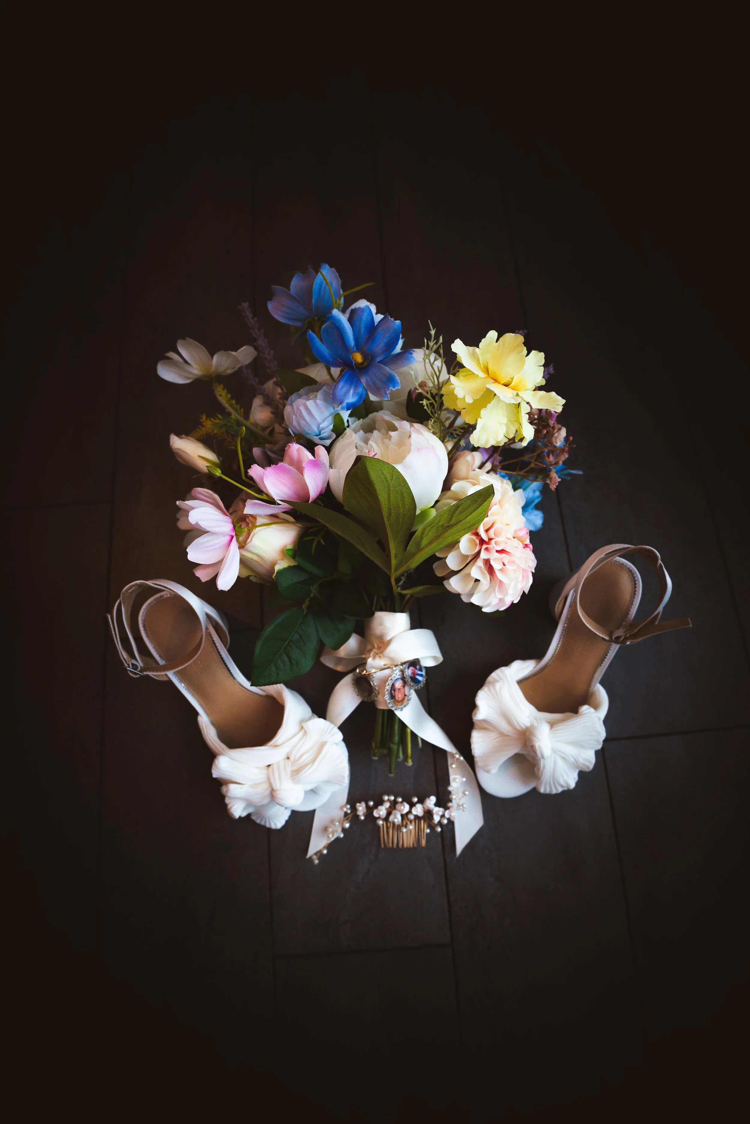 A bouquet of colorful flowers, including blue, yellow, pink, and white blooms, placed on a dark wooden floor, with a pair of white high-heeled shoes with bows and ribbon accents below.