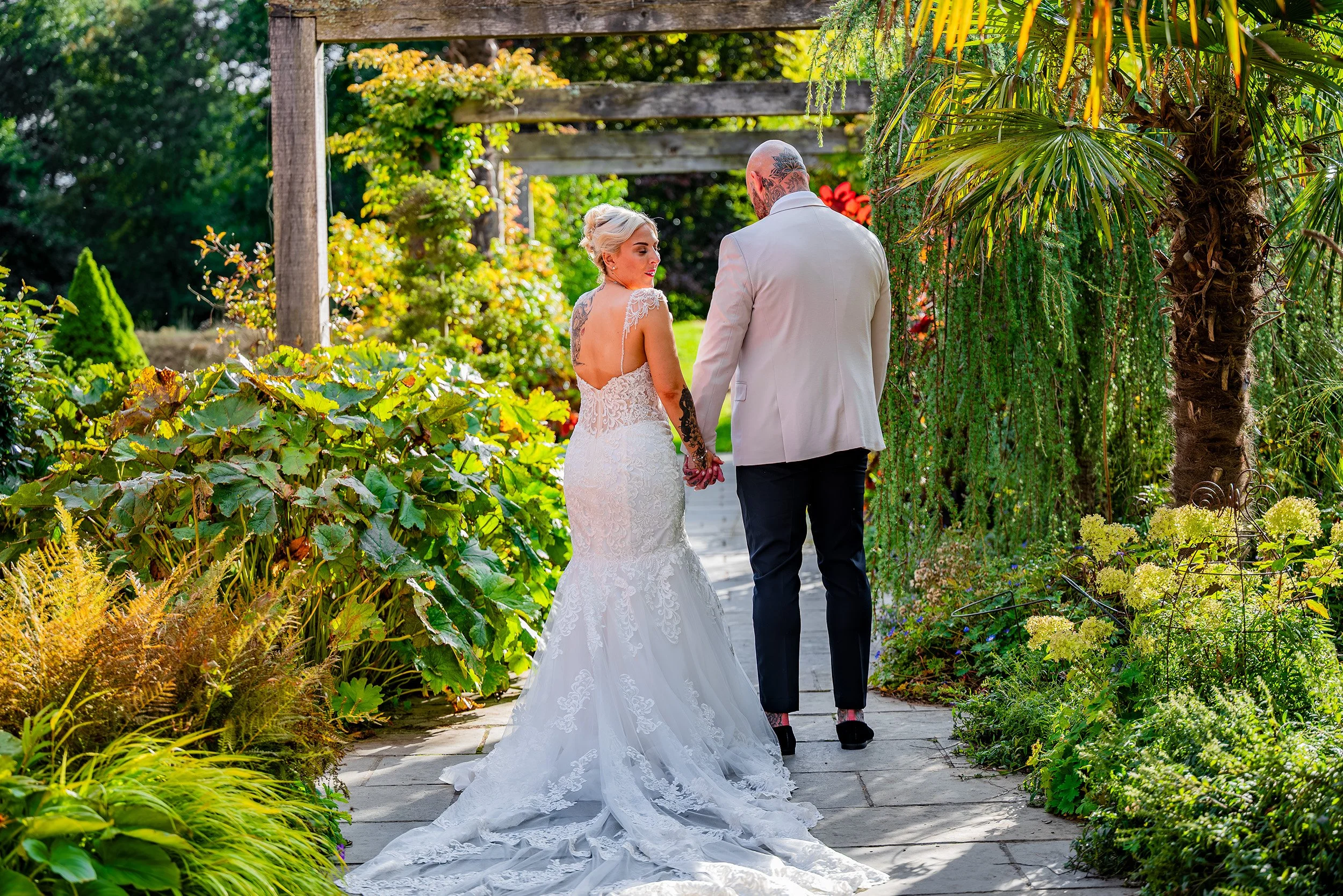 A bride and groom walking hand in hand through a lush garden pathway on their wedding day, surrounded by greenery and flowering plants.