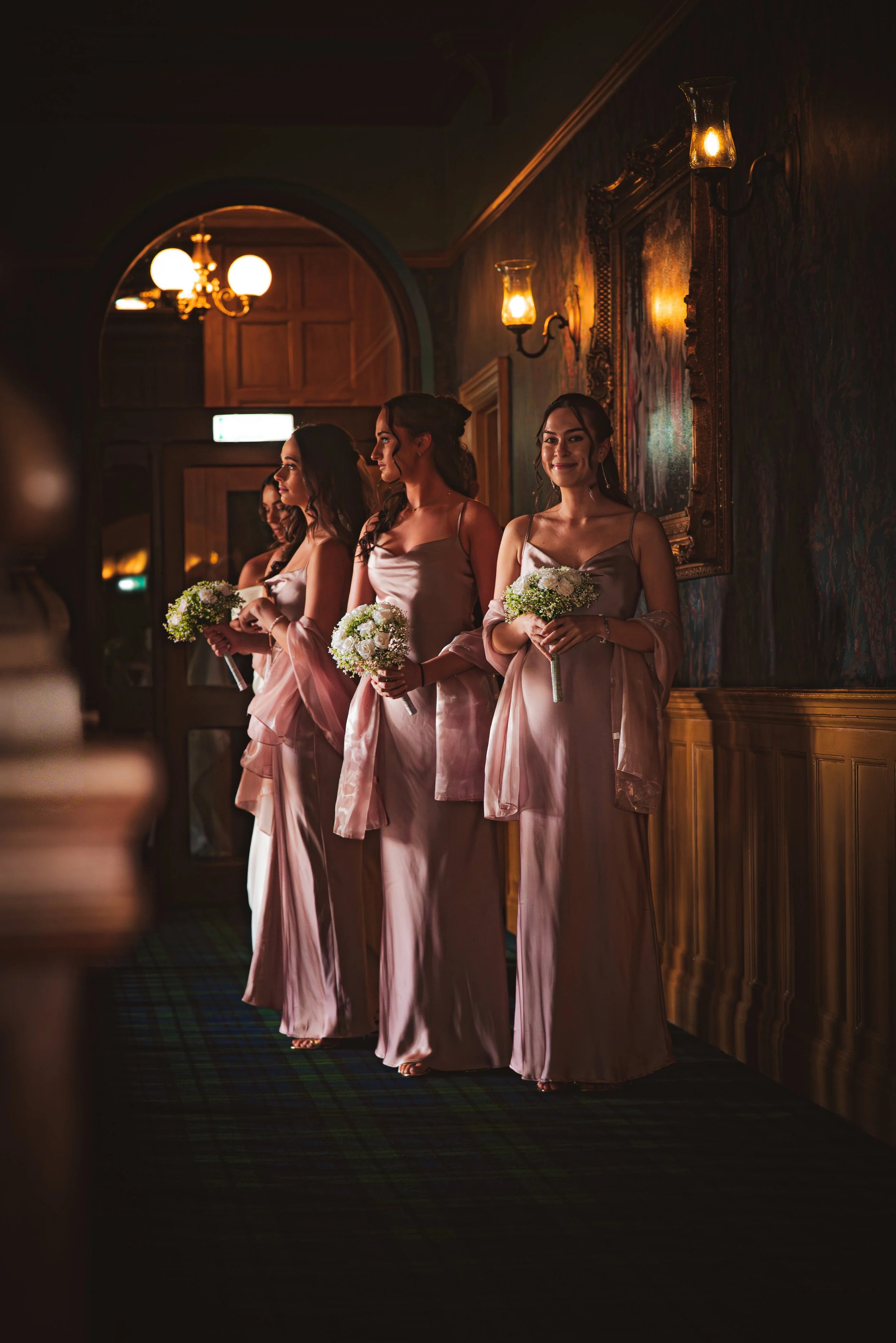 Four women dressed in pink satin gowns holding bouquets, standing in a dimly lit hallway with vintage wall sconces and a large framed mirror.