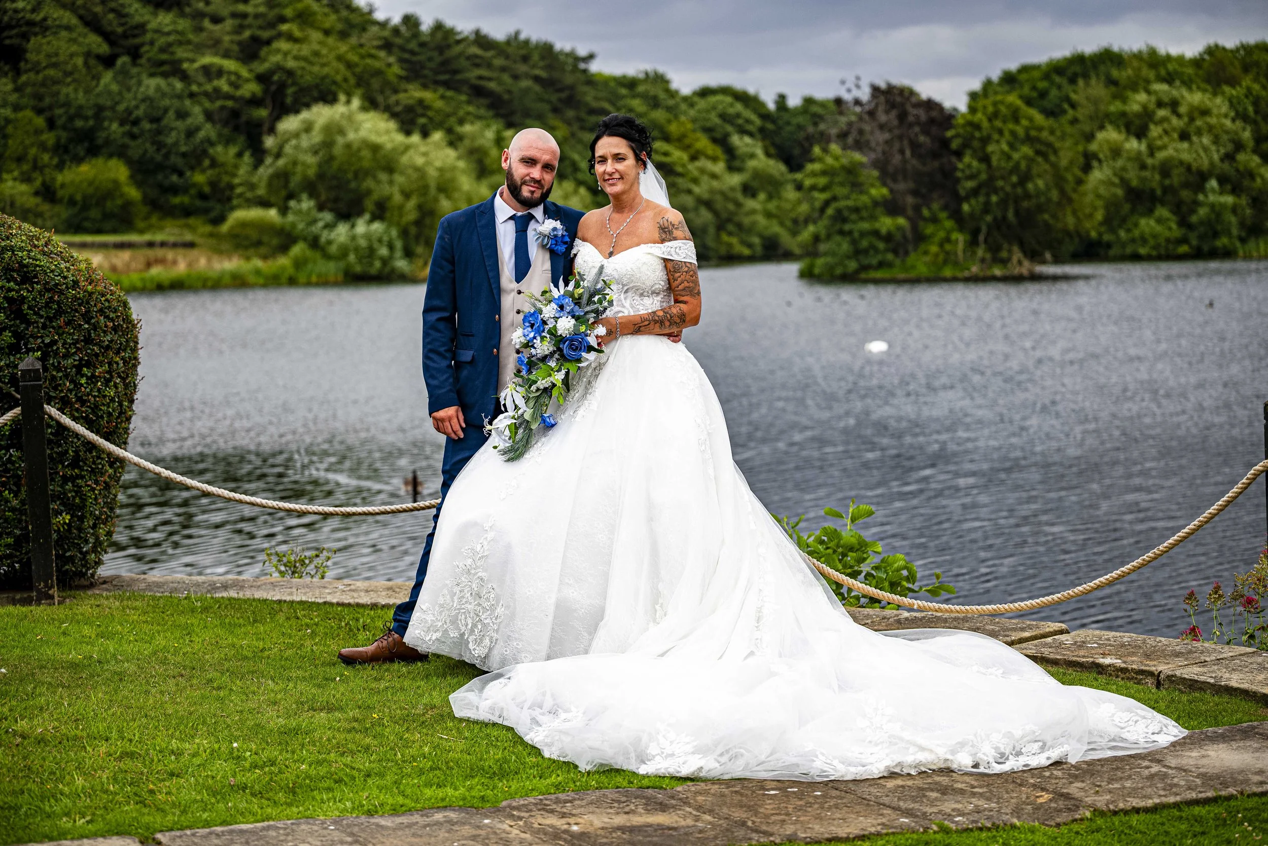 Bride in white wedding dress and groom in blue suit standing by lake with greenery and trees in background.