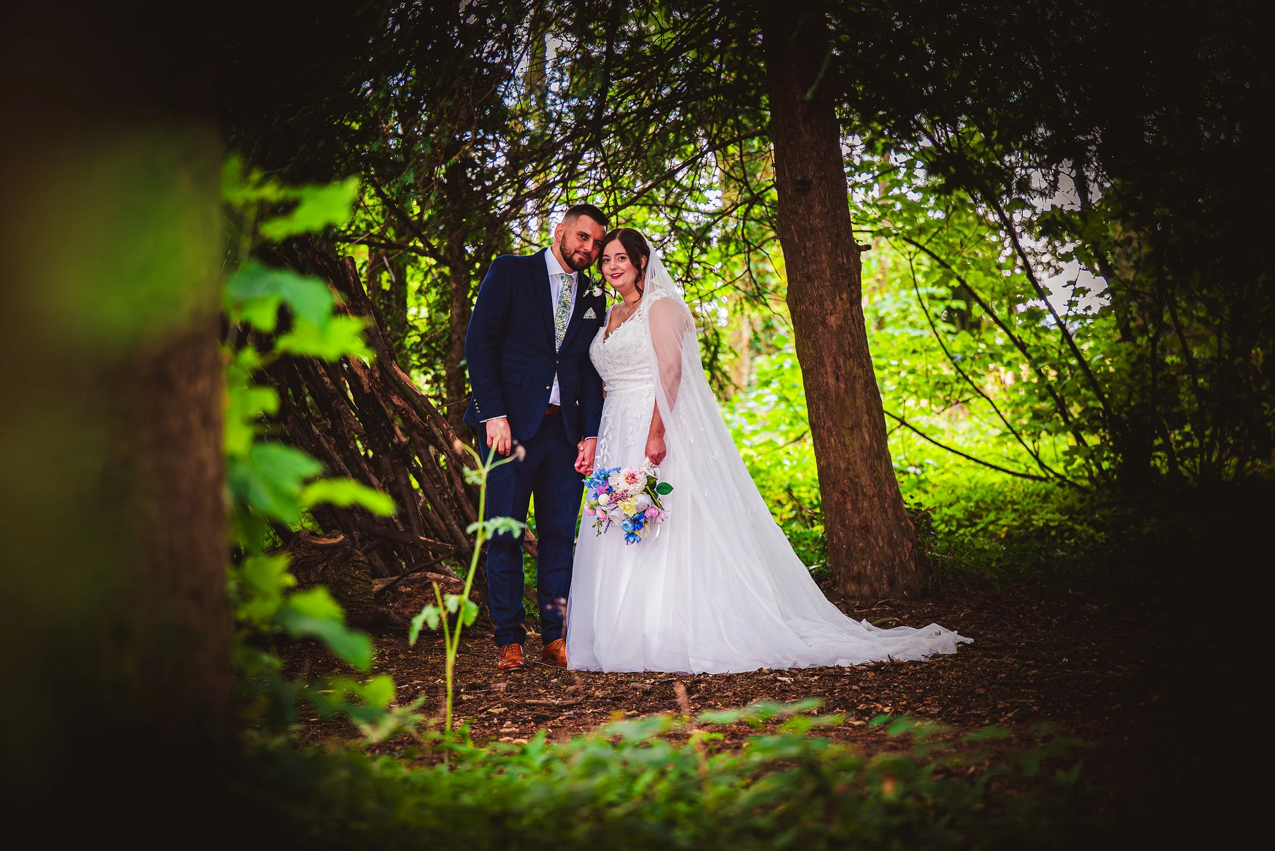 A bride and groom standing hand in hand in a lush green forest during their wedding, with the bride in a white wedding gown holding a bouquet of colorful flowers and the groom in a dark blue suit.