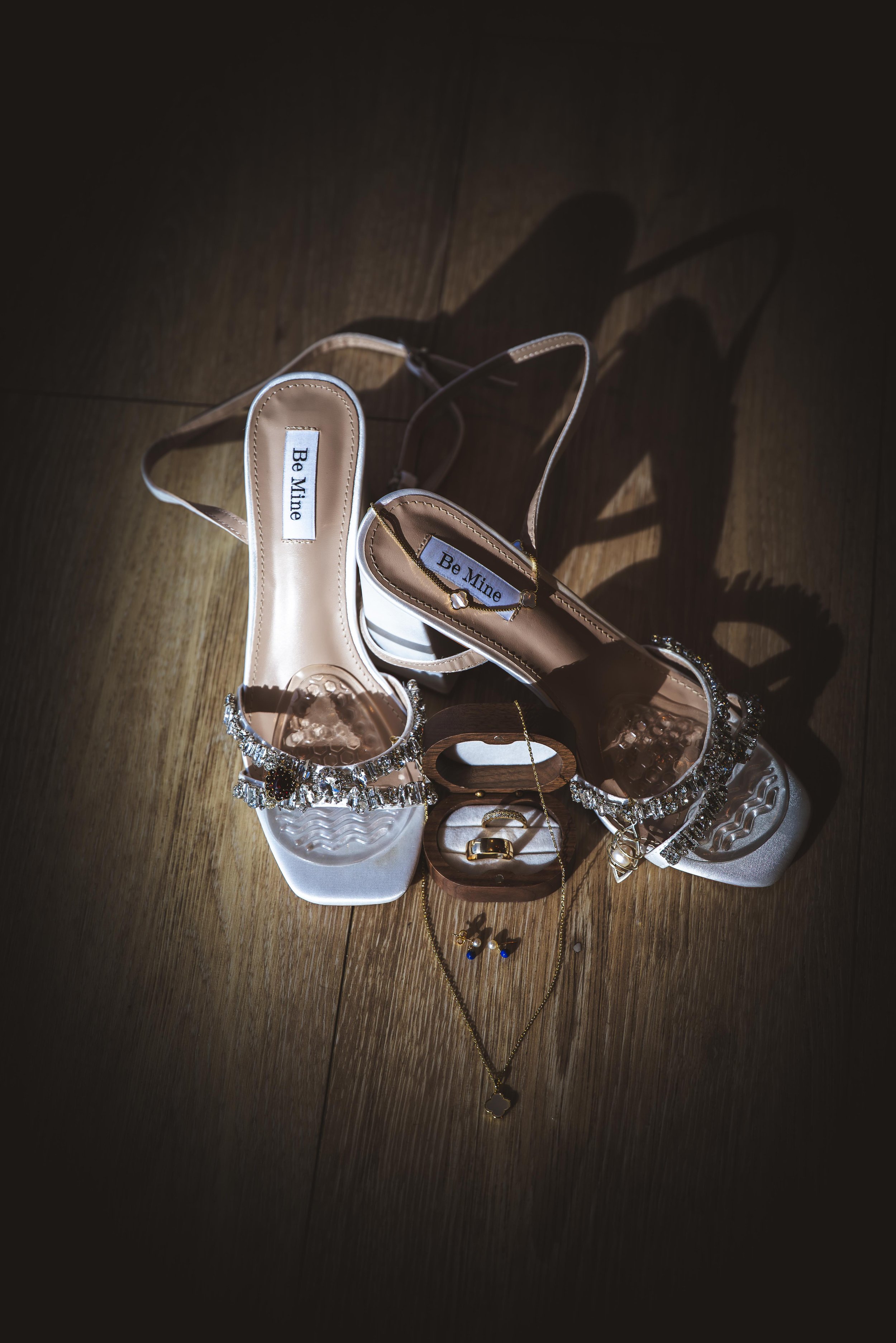 A pair of white high-heeled sandals with rhinestone embellishments, a small jewelry box with a gold ring, a gold necklace with a pendant, and a pair of earrings, all arranged on a wooden floor with a spotlight creating shadows.