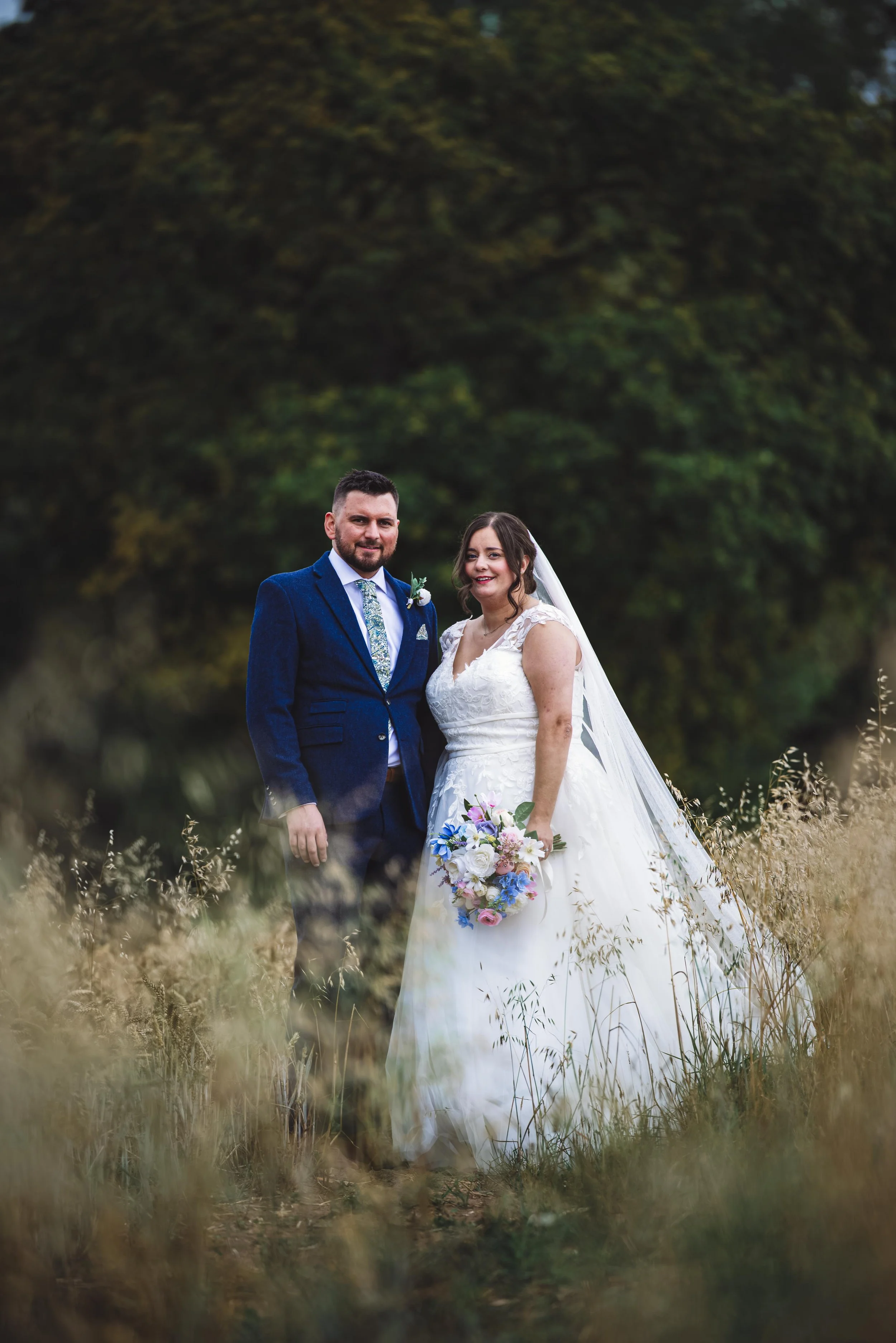 A bride and groom standing in a field with tall grass, with trees in the background, on their wedding day.
