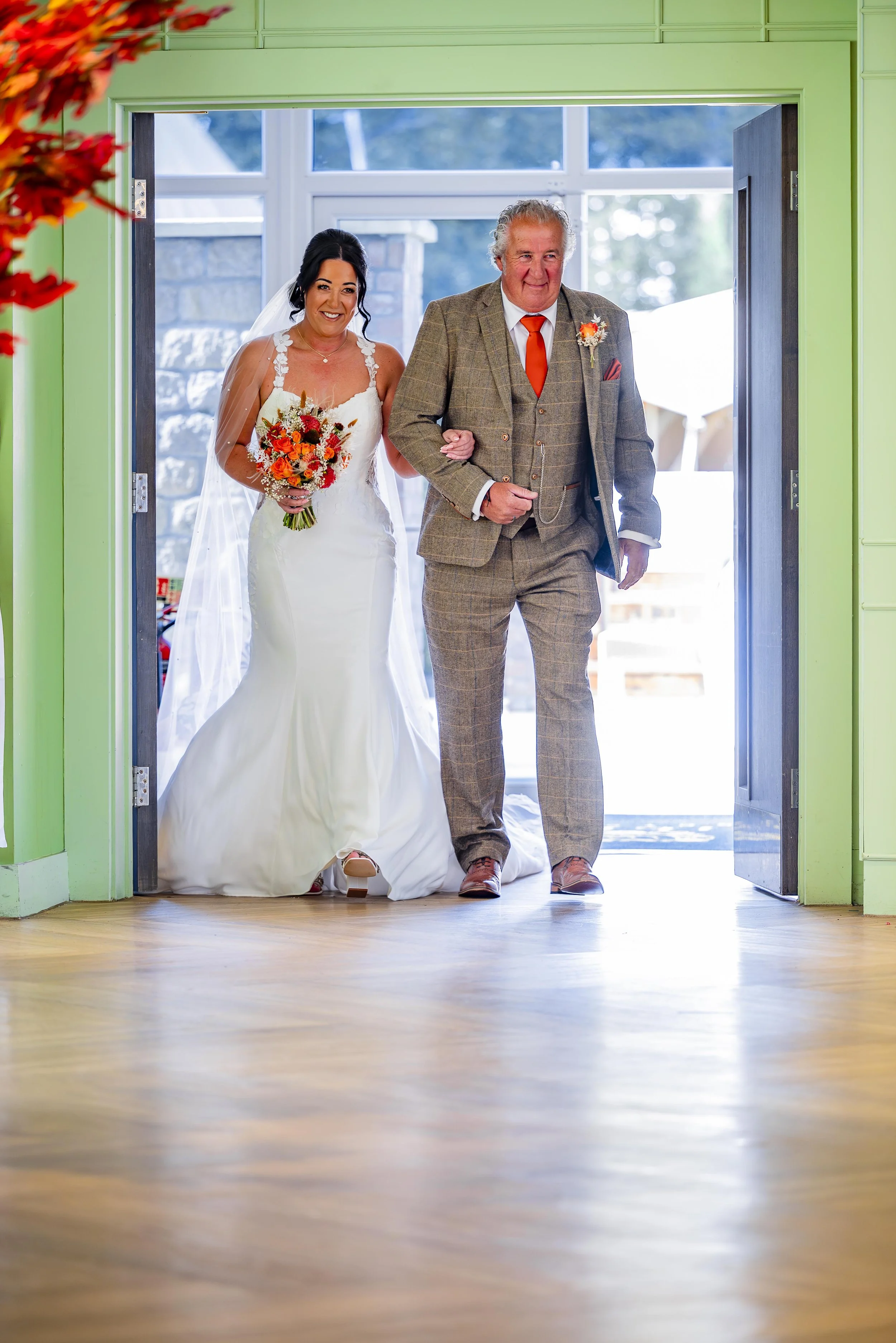 Bride walking down the aisle with her father at a wedding, holding a bouquet of orange and red flowers, inside a room with green walls.
