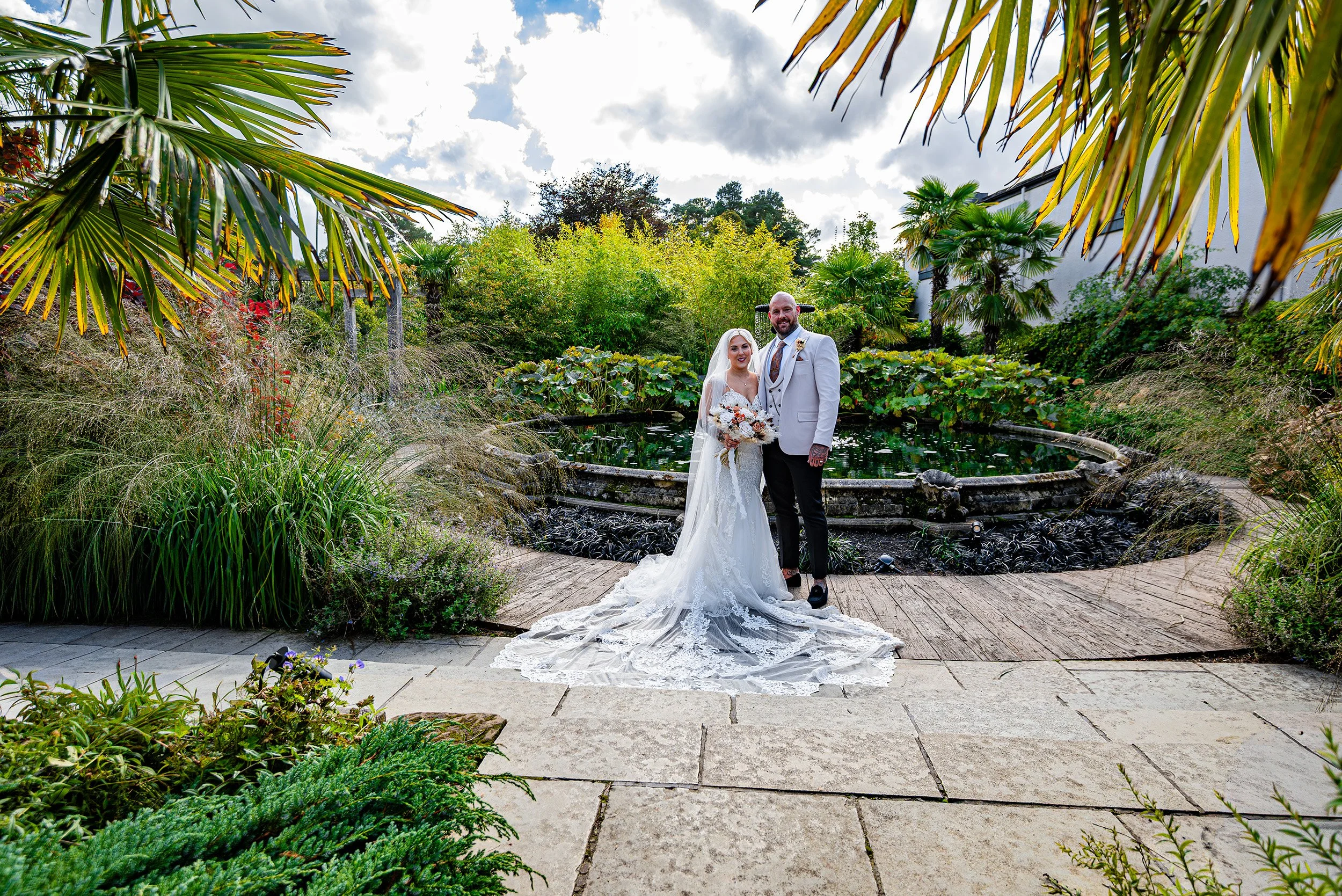 A bride and groom standing together outdoors in front of a pond, surrounded by lush greenery and tropical plants, under a cloudy sky.