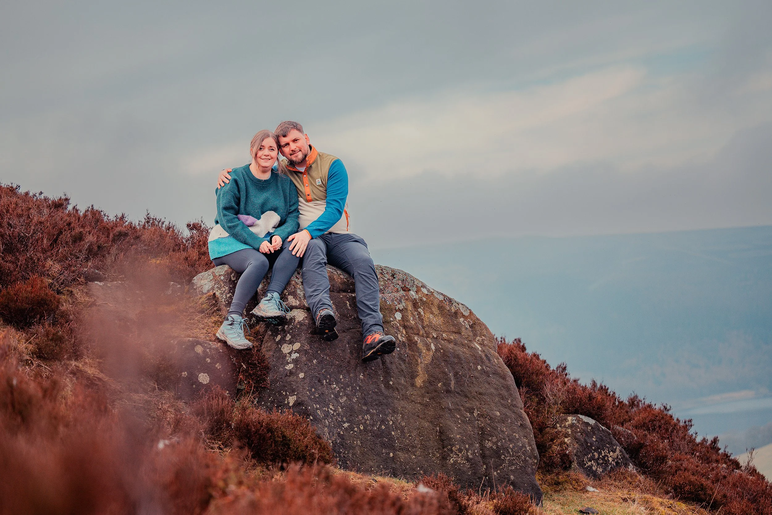 A smiling couple sitting on a large rock on a hillside with brownish shrubbery, outdoors during daytime, under a cloudy sky.
