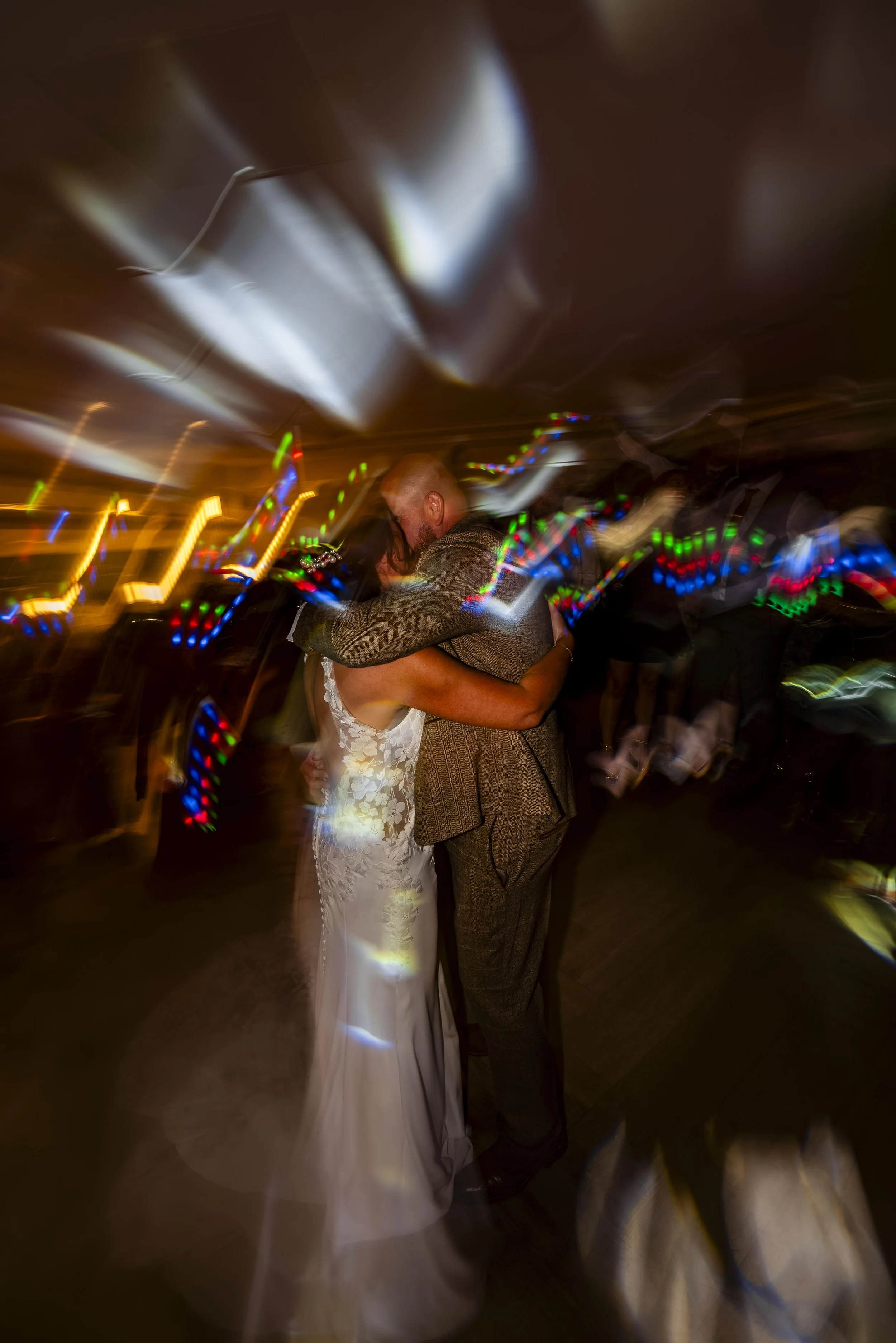 A man and woman embracing and dancing at what appears to be a wedding reception, with colorful lights and motion blur creating an energetic atmosphere.
