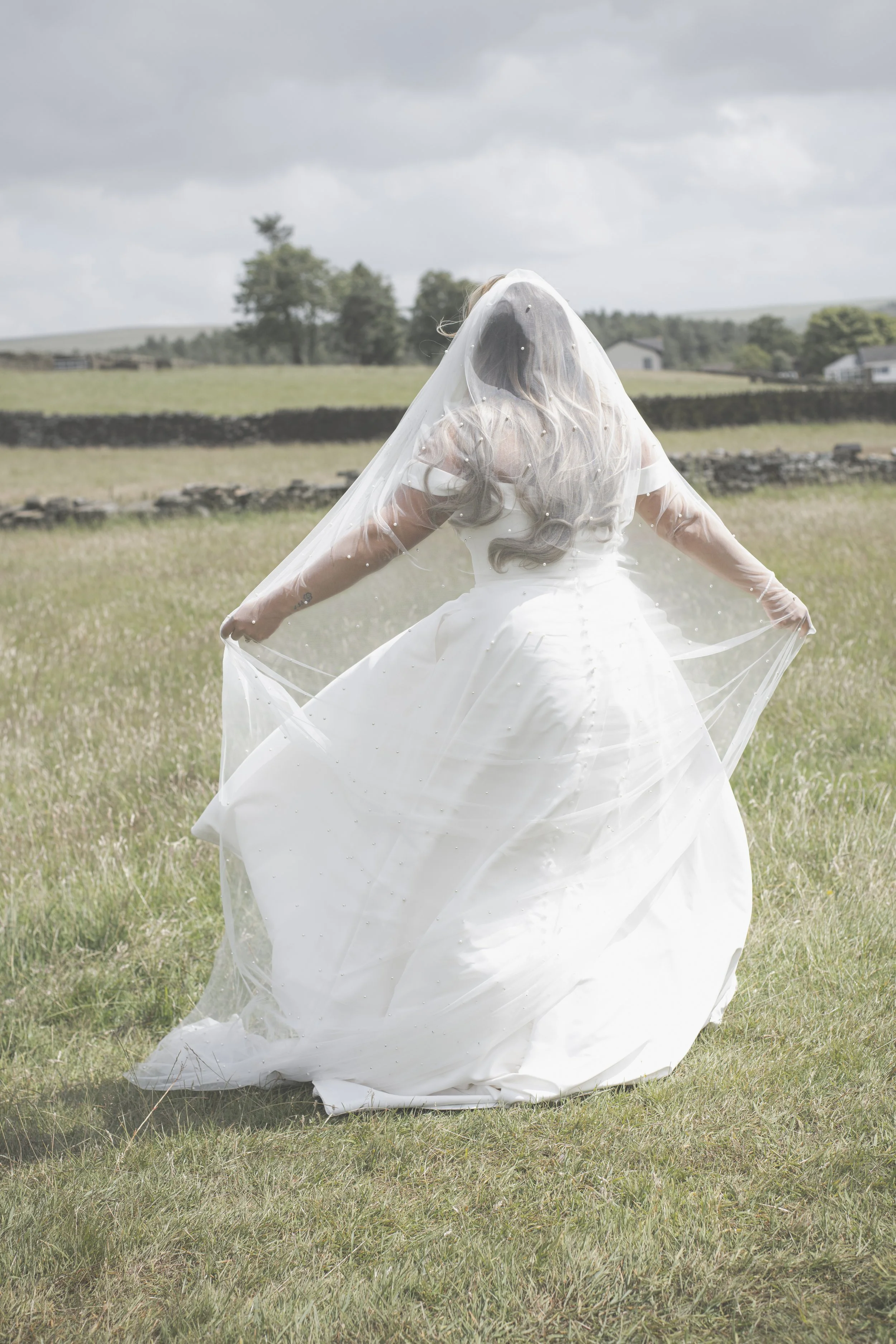A woman in a white wedding dress and veil standing in an open grassy field with her back facing the camera, holding the sides of her dress and veil.