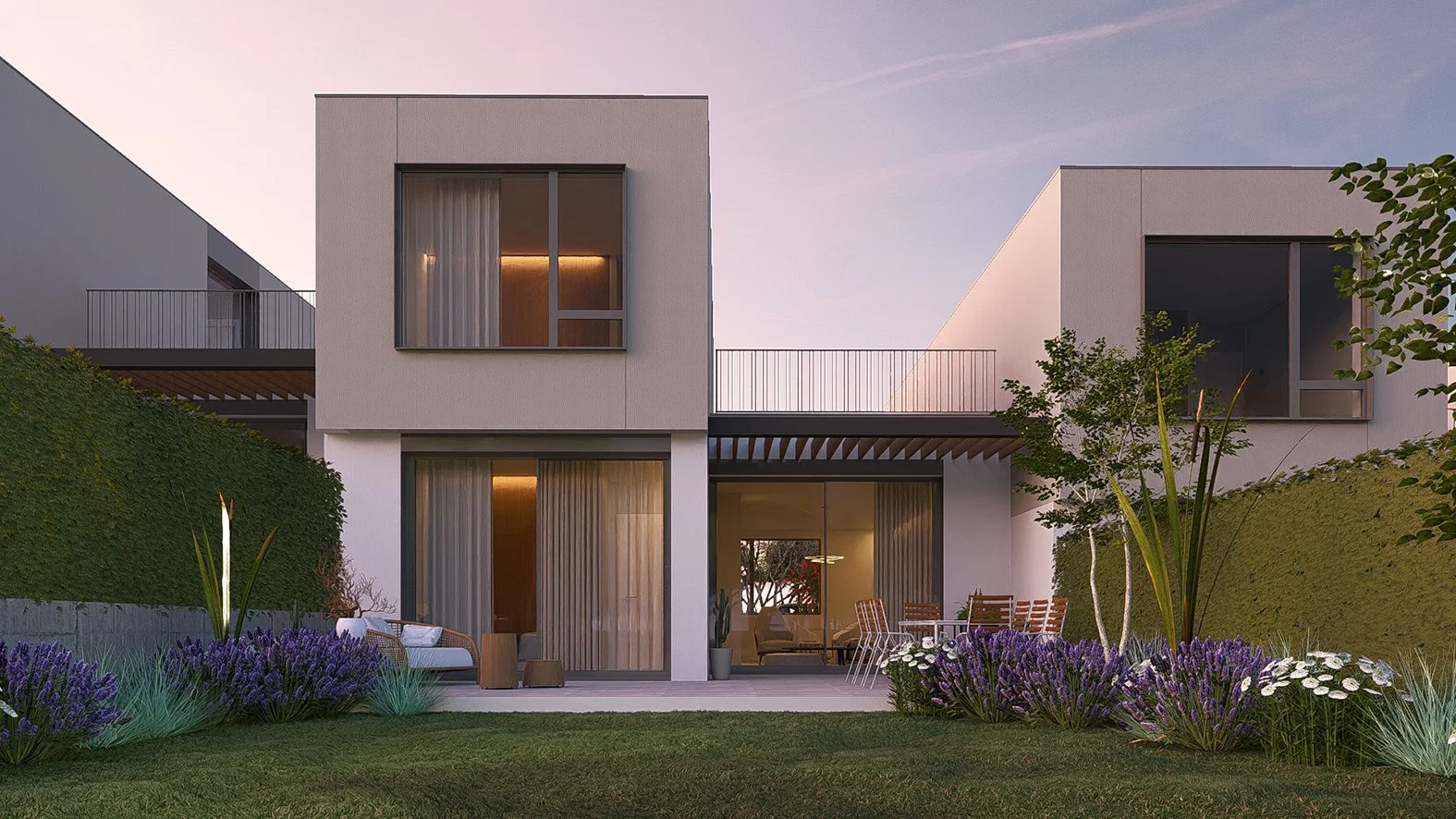 Vista de una casa moderna de dos niveles con paredes blancas, ventanas grandes con cortinas, y un jardín con plantas y flores moradas, durante el atardecer.