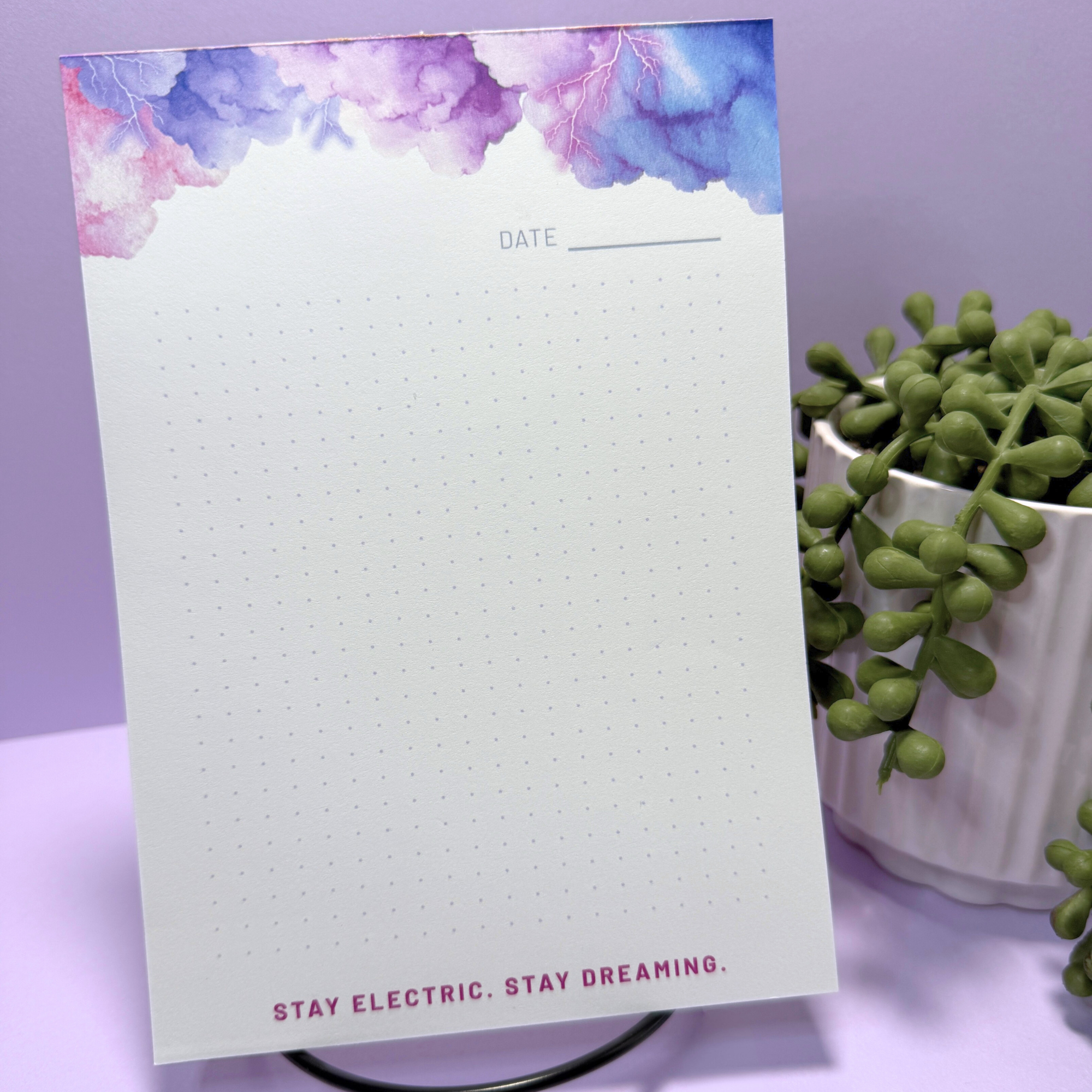 A blank notepad with a purple and pink cloud and lightning storm watercolor border at the top, and the phrase "Stay electric. Stay dreaming." at the bottom, placed next to a potted succulent plant with chunky green leaves.