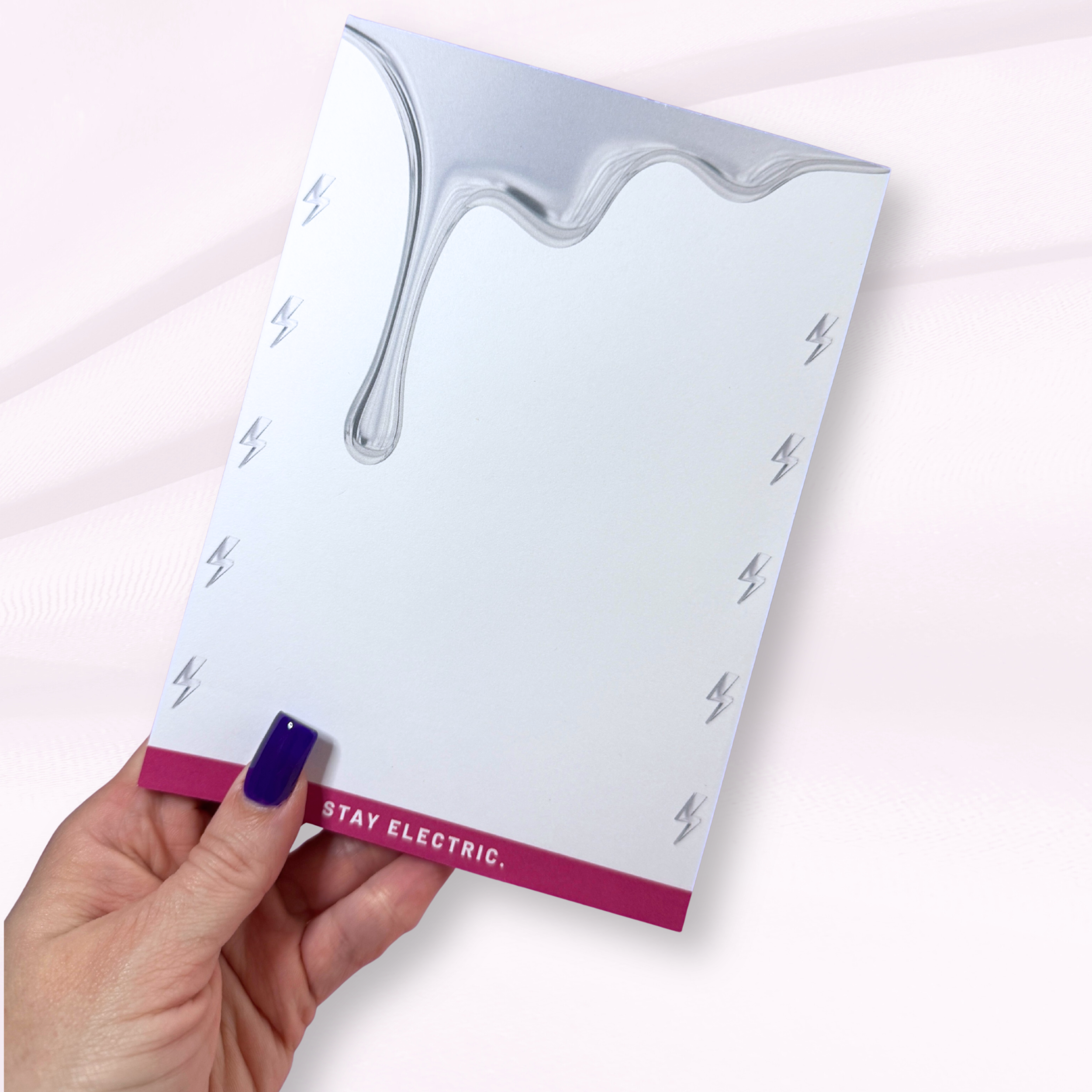 A hand holding an electrical safety poster with a silver metallic finish and a pink bottom border that reads 'STAY ELECTRIC.' The poster features a melting metallic design at the top and small lightning bolt symbols along the sides.}