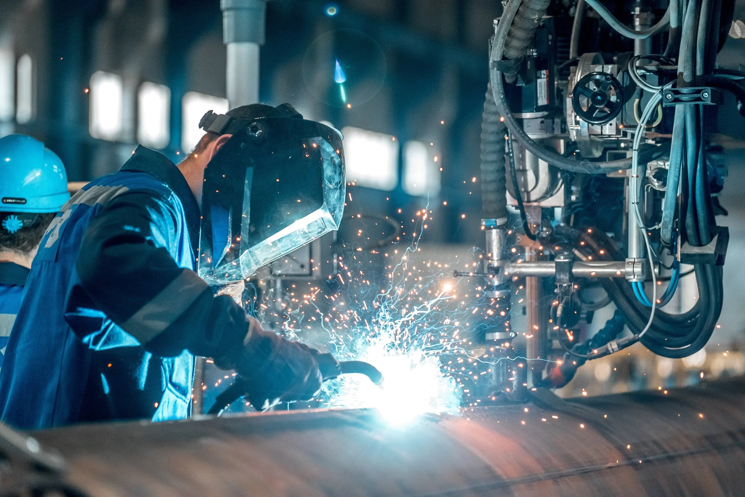 Welder wearing a protective helmet and gloves welding a metal pipe, with sparks flying in an industrial workshop.
