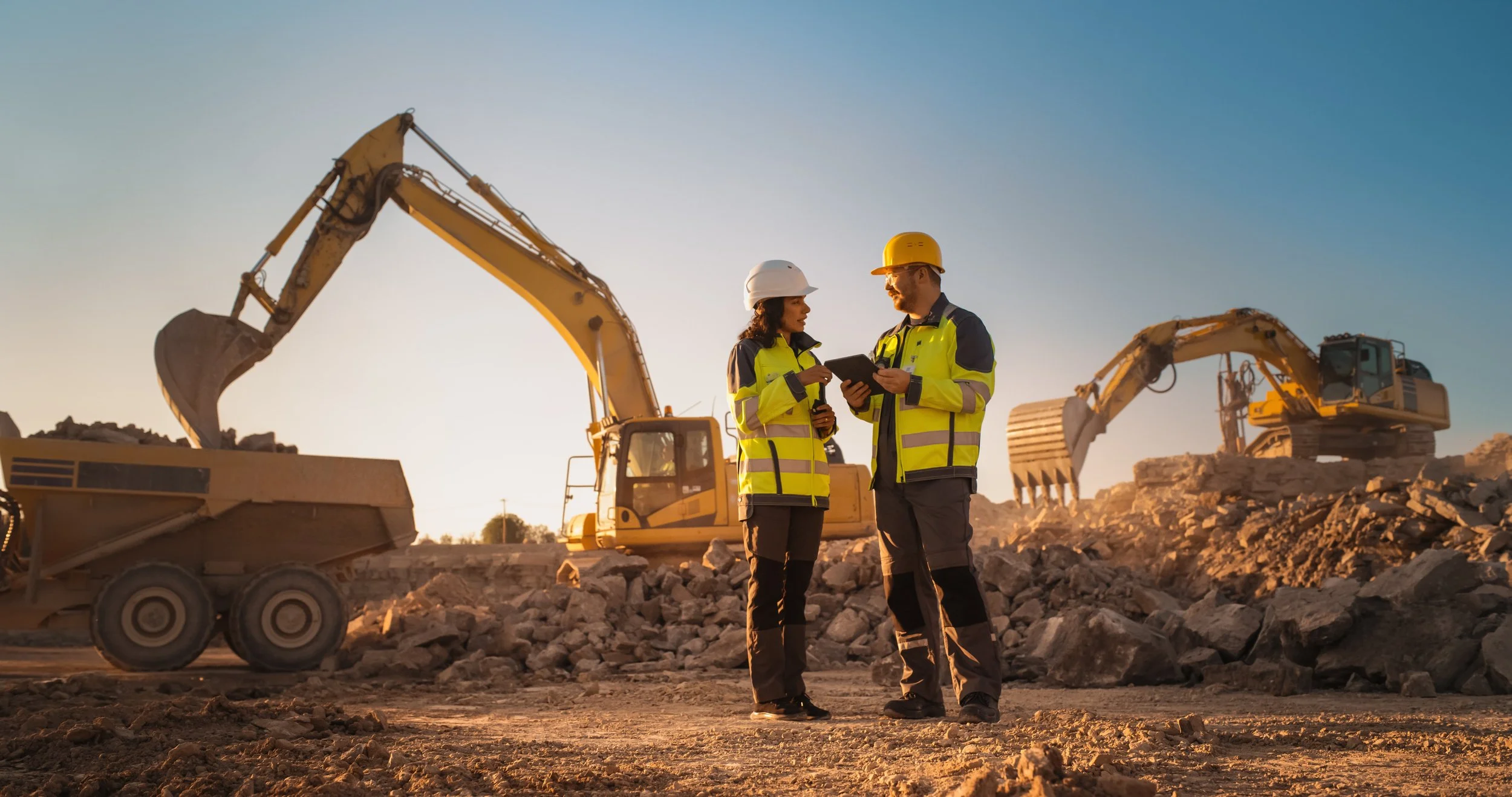 Two construction workers in safety gear, including hard hats and reflective jackets, standing in a construction site with excavators and rocks in the background, discussing work plans during sunset.