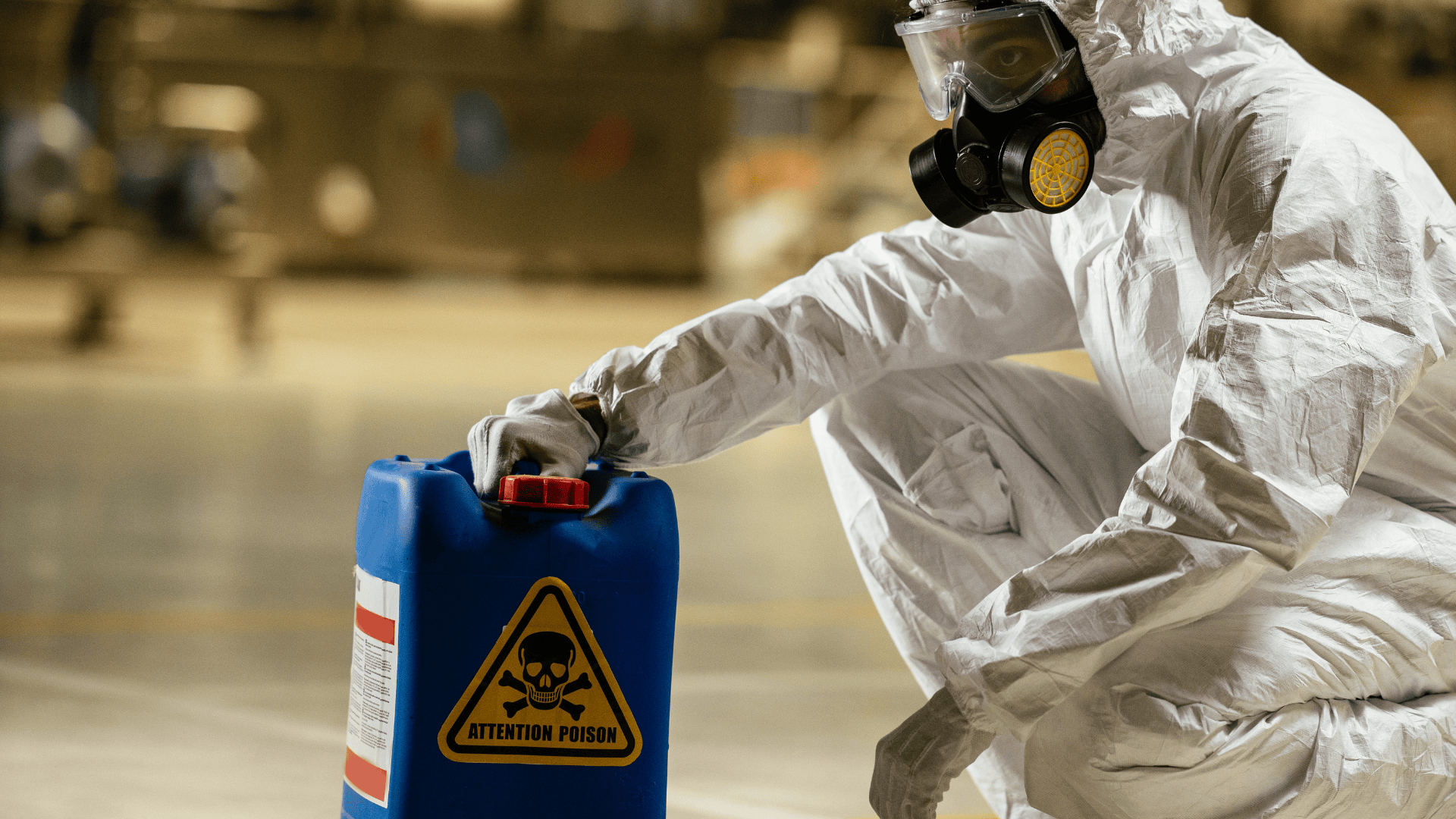 Person dressed in white hazmat suit with respirator mask, kneeling and holding a blue container with a poison label.