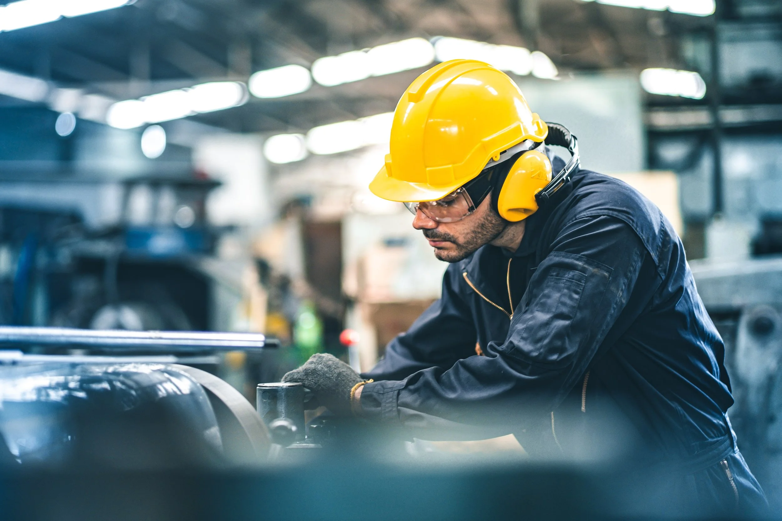 A male worker in a manufacturing or industrial setting wearing a yellow hard hat, safety goggles, and ear protection, working with machinery.