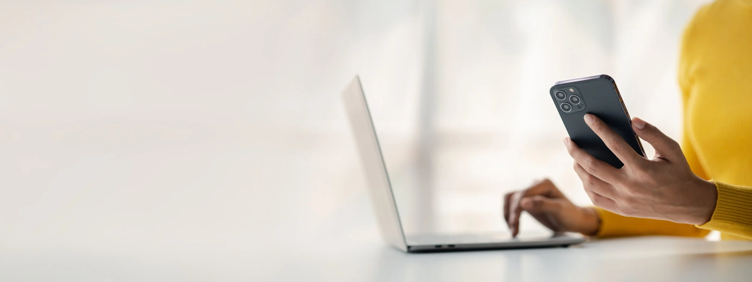 Person in a yellow sweater holding a smartphone while working on a laptop at a white desk.