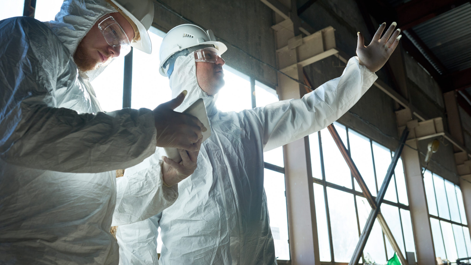 Two construction workers in white protective suits, helmets, and safety glasses inside a building with large windows, one pointing outward and the other taking notes on a mobile device.