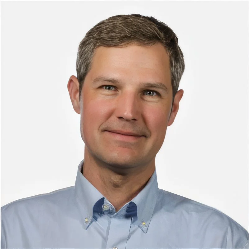 A smiling man with short brown hair wearing a light blue collared shirt against a plain white background.