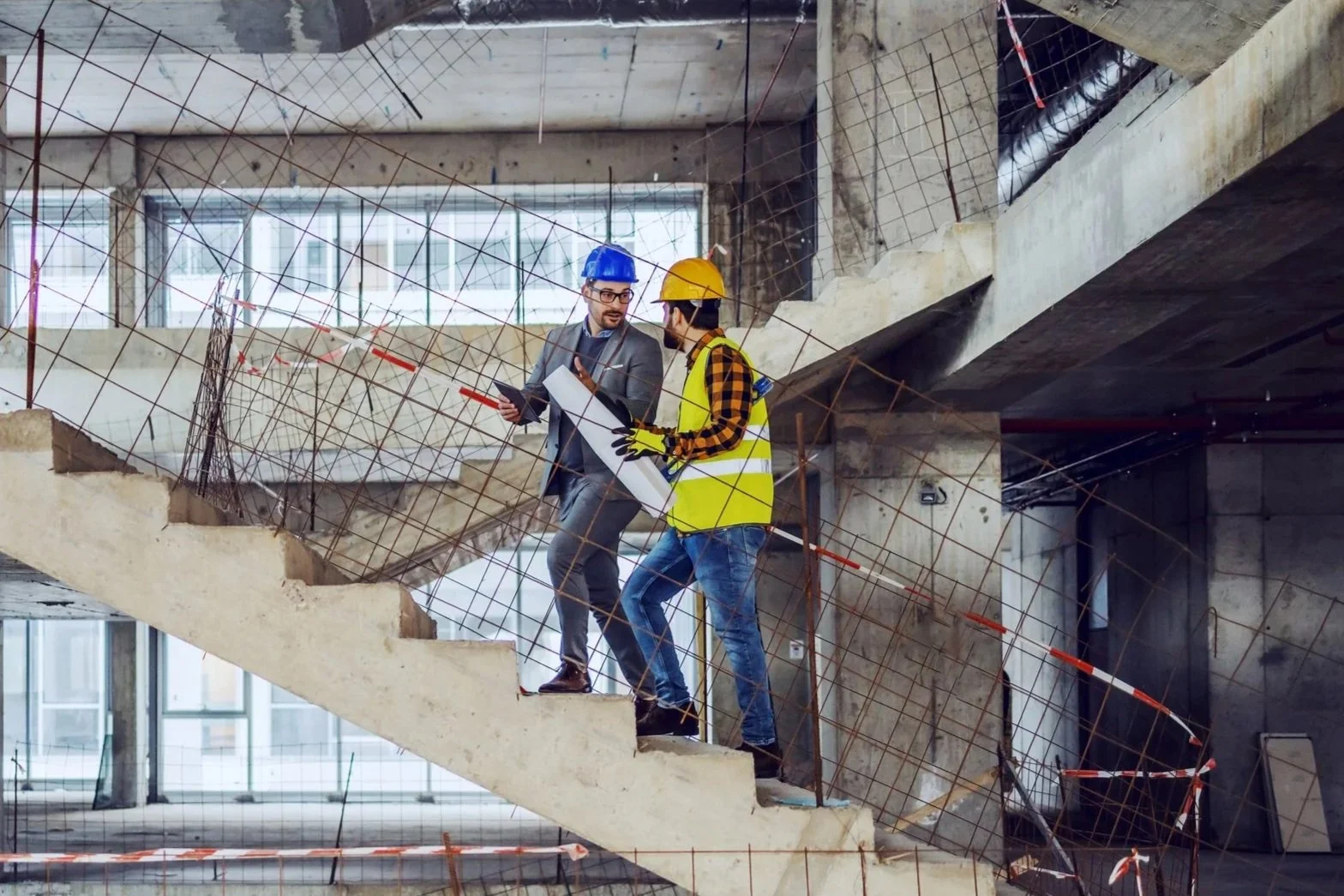 Two construction workers, one in a suit and safety helmet, and the other in a yellow safety vest and helmet, discussing plans on a construction site stairway.