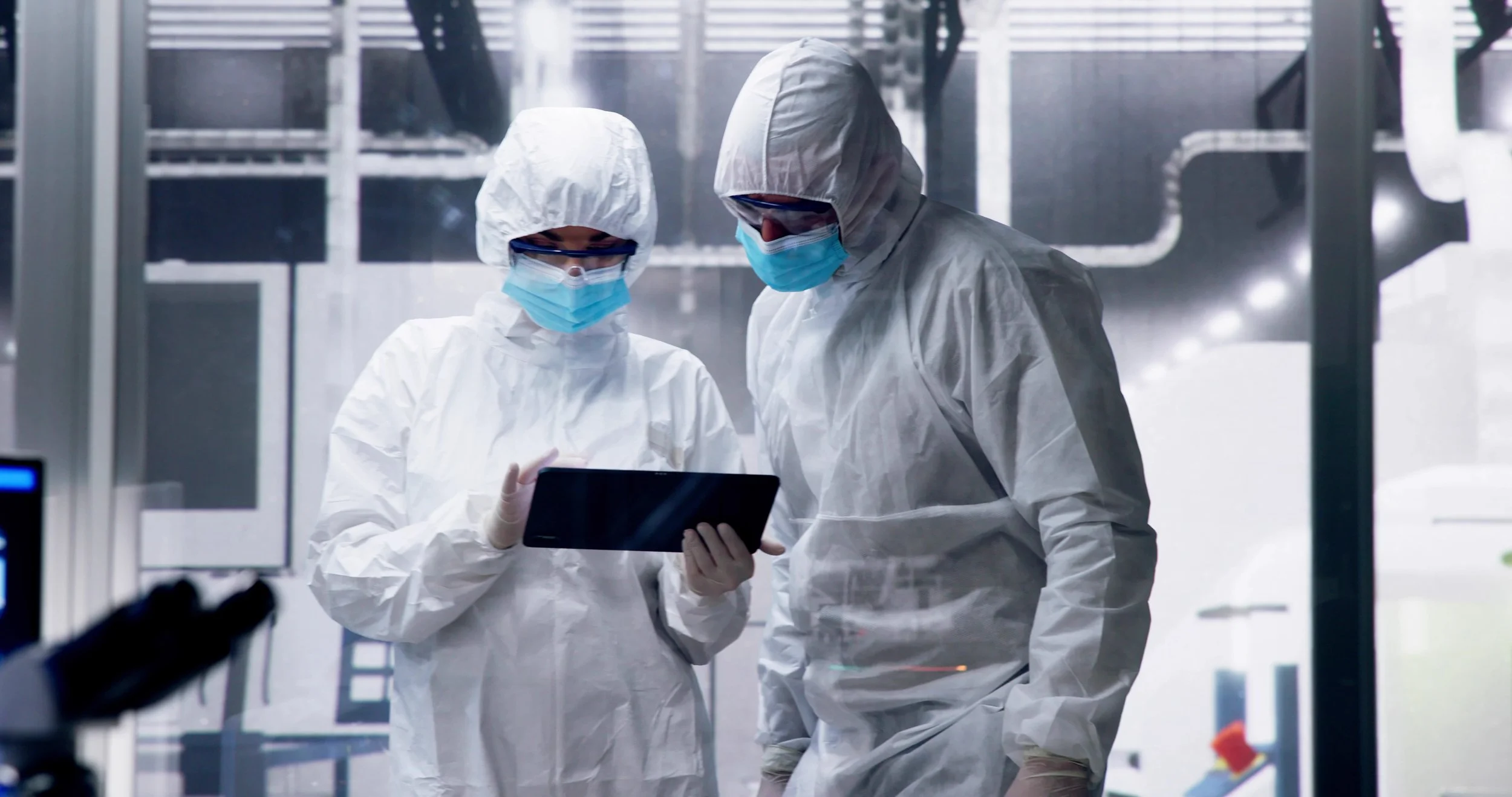 Two scientists in full protective gear, including masks and goggles, examining a tablet in a high-tech laboratory.