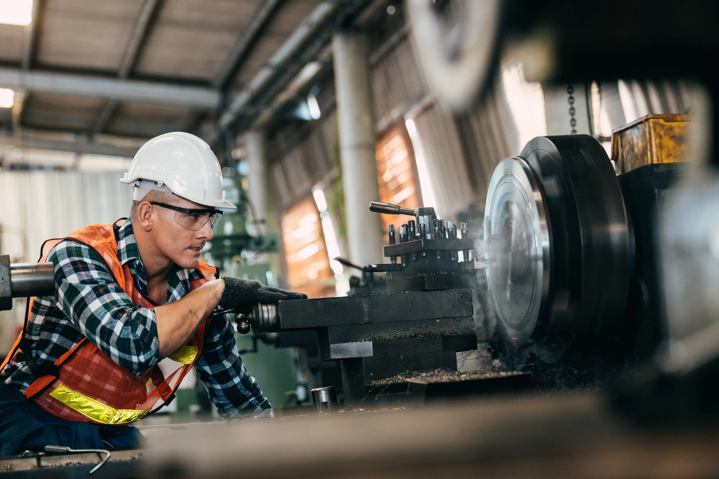 A factory worker wearing safety glasses, a white hard hat, a plaid shirt, and an orange safety vest operates a large machine, which is spinning a metal disc, inside an industrial workshop.