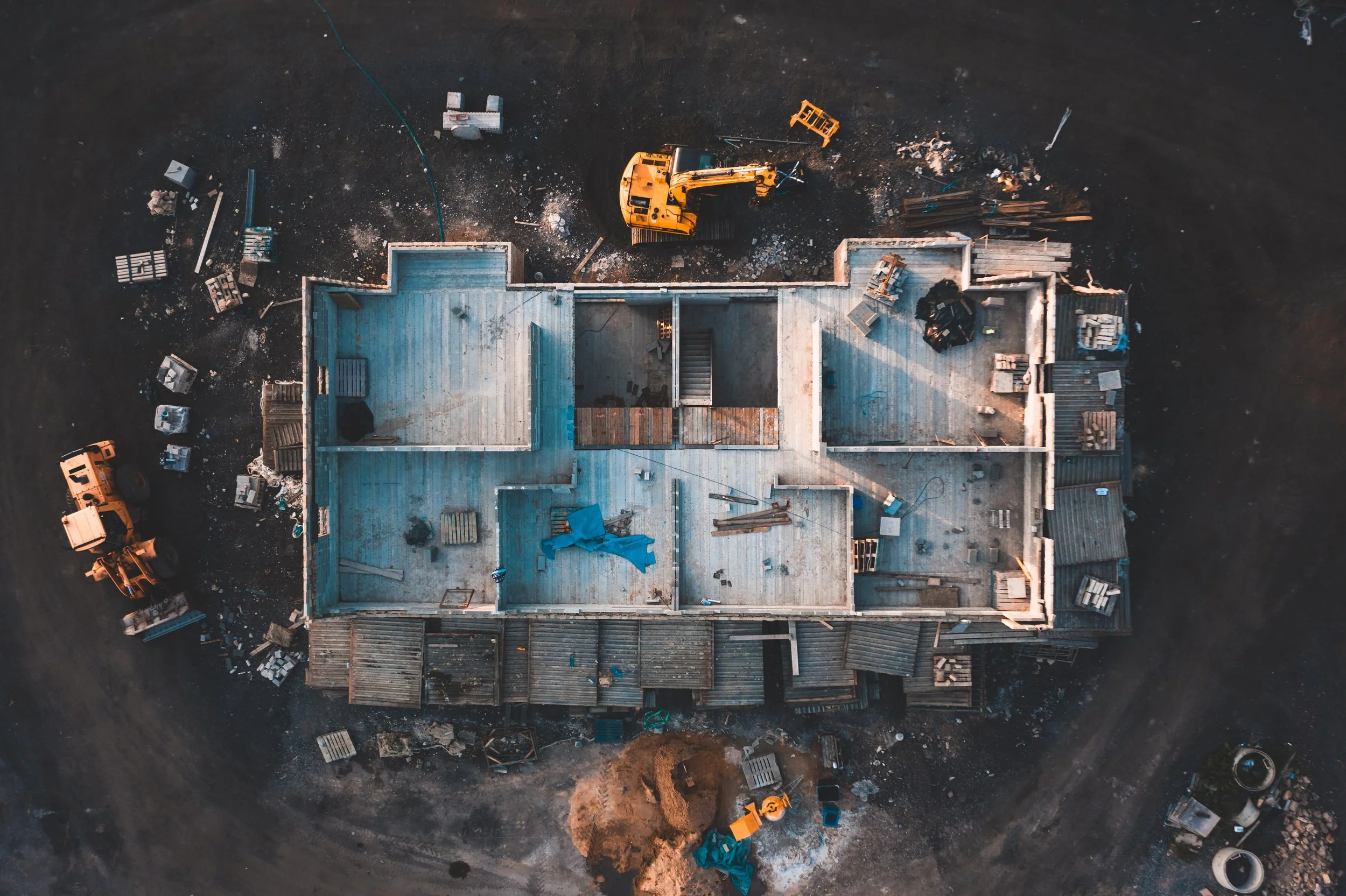 An aerial view of a house under construction with a yellow excavator and construction materials scattered around.