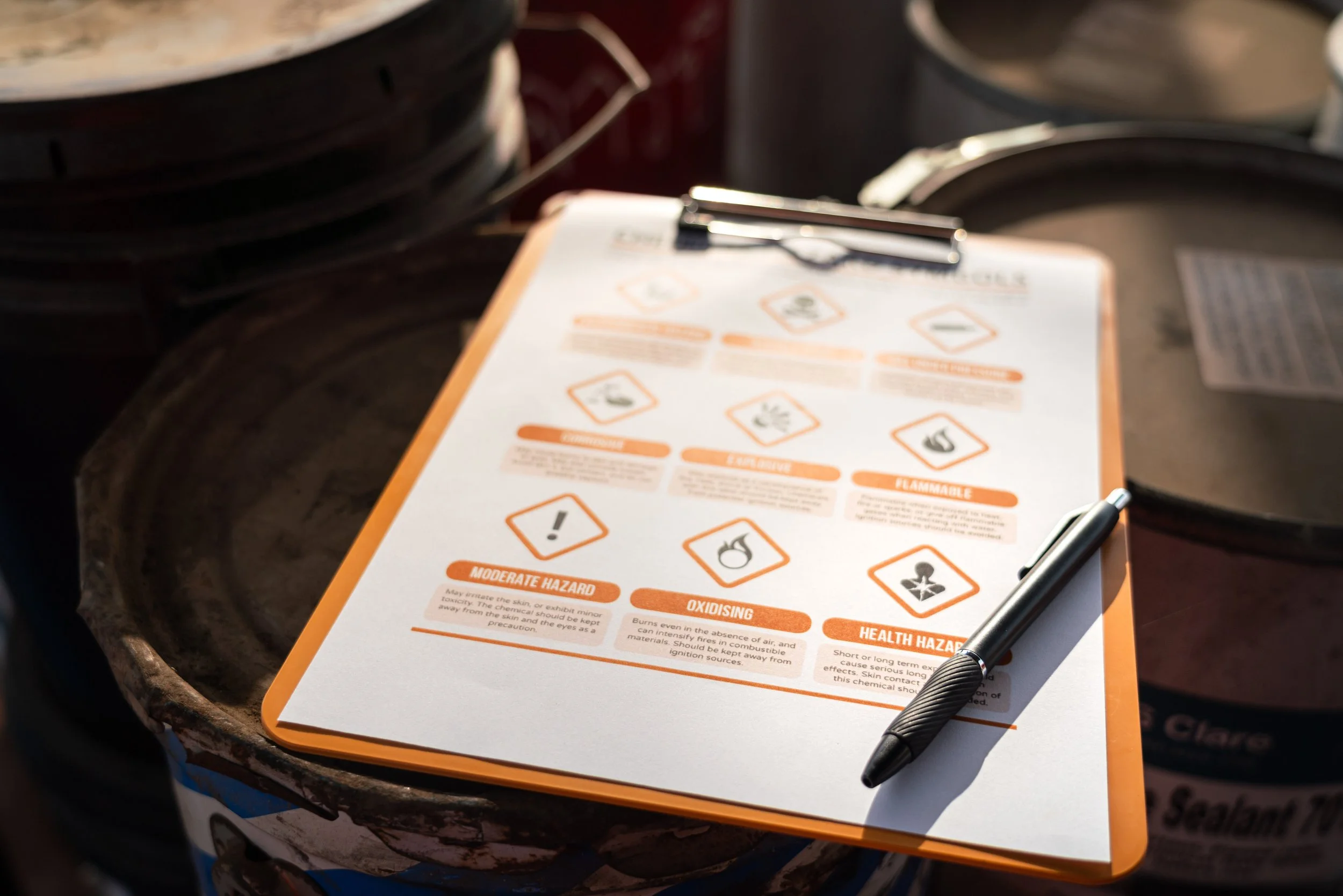 A clipboard with a safety data sheet on hazardous chemicals, placed on top of large containers or drums with warning labels.