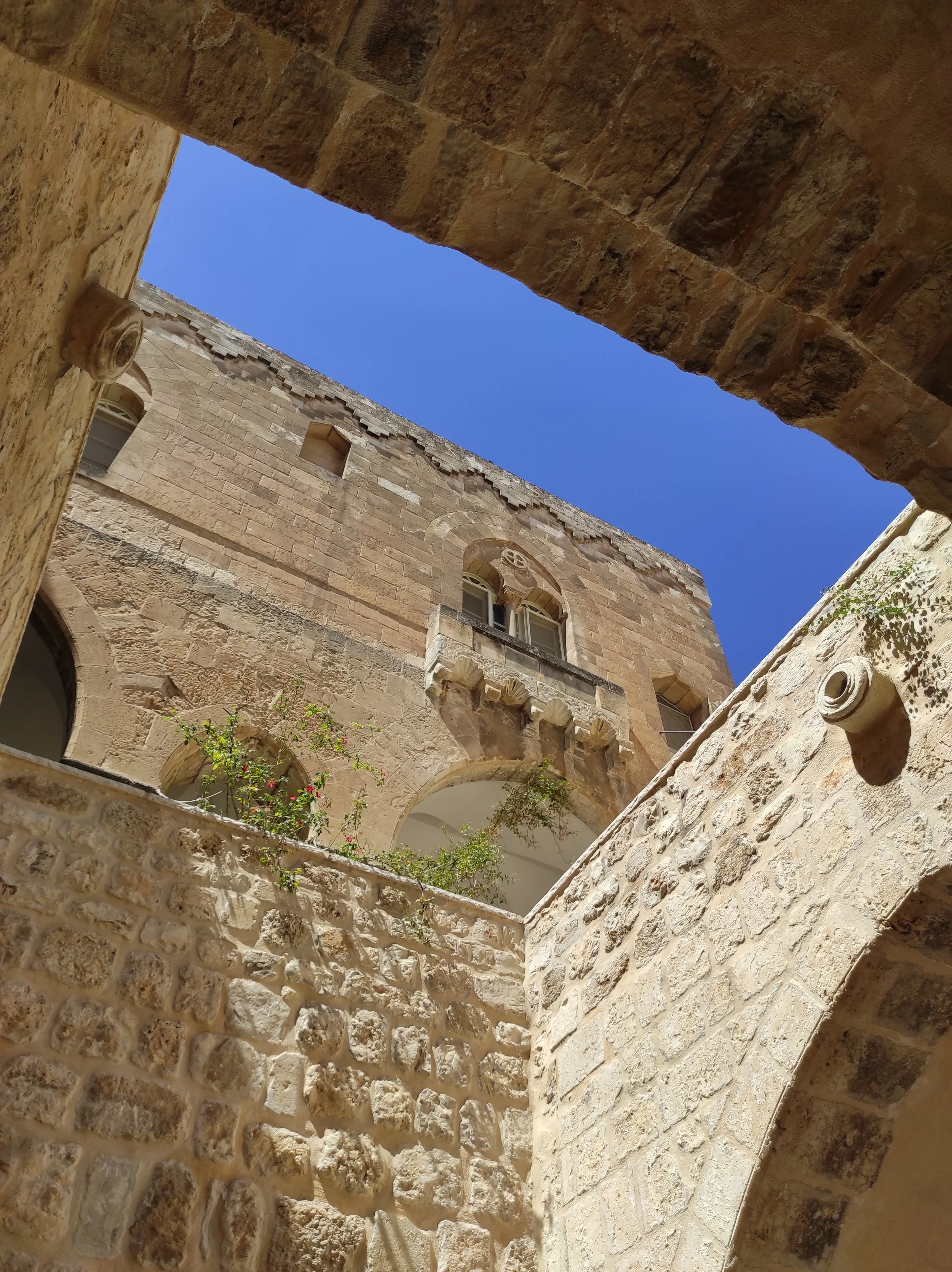 View looking up through a stone courtyard at a historic stone building with arched windows, stone balconies, and small plants, against a bright blue sky.
