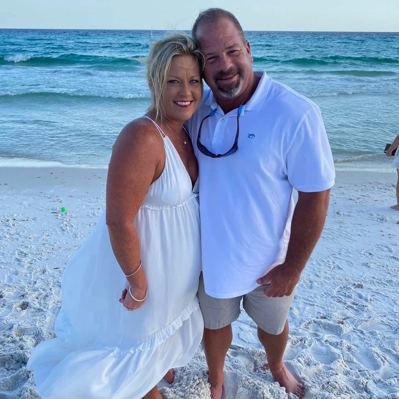 Jesse Grant is standing close to his wife on a beach with white sand and ocean waves in the background. The woman is wearing a white dress and the man is wearing a white polo shirt and shorts.
