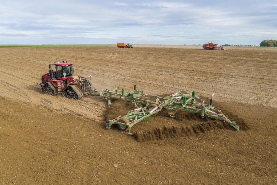 A Case red tractor pulling a KELLY Tillage Diamond Harrow across a large open field with two additional tractors in the background, working on the expansive farmland under a cloudy sky.