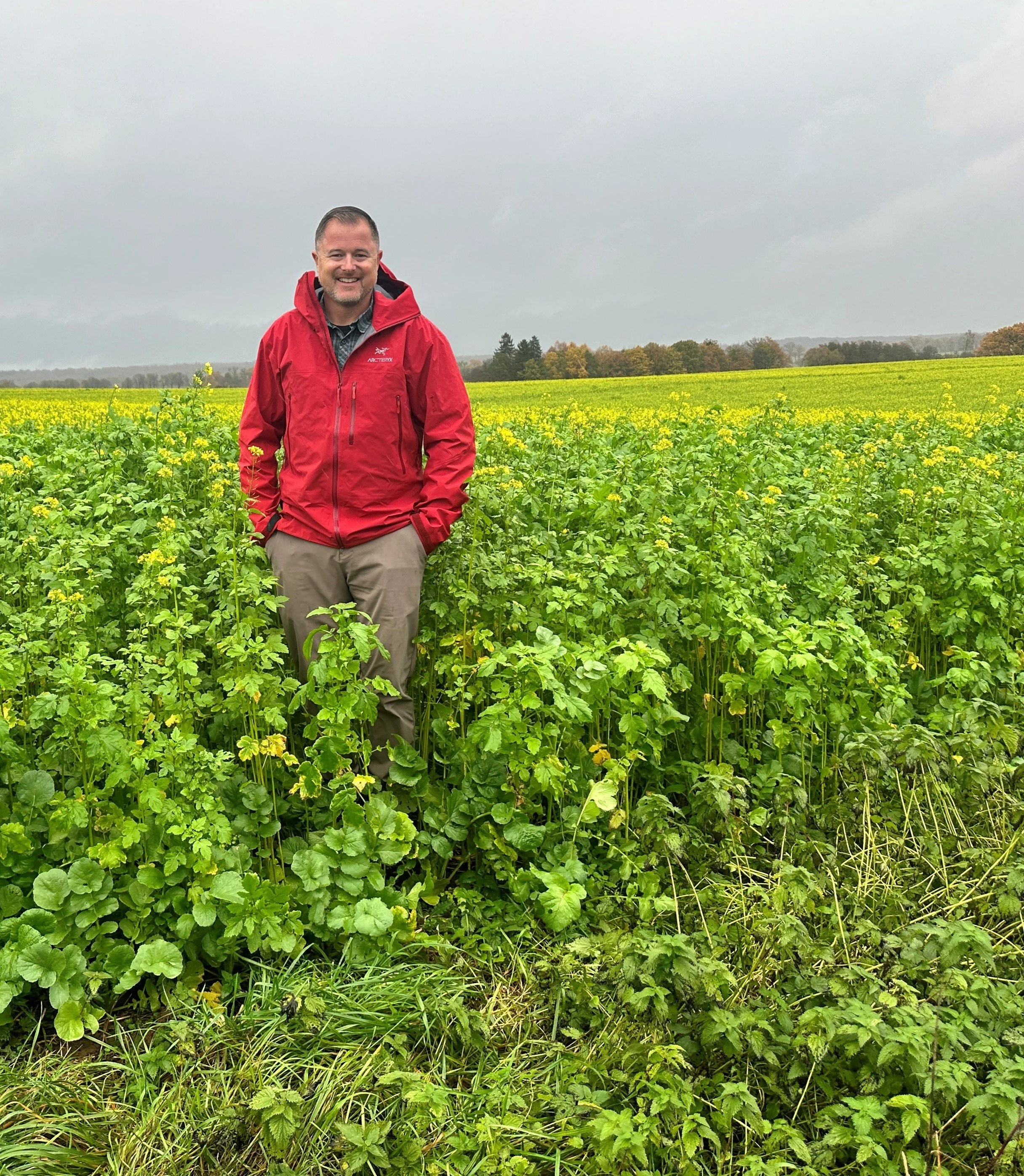 A man standing in a green field of plants, wearing a red jacket and smiling.