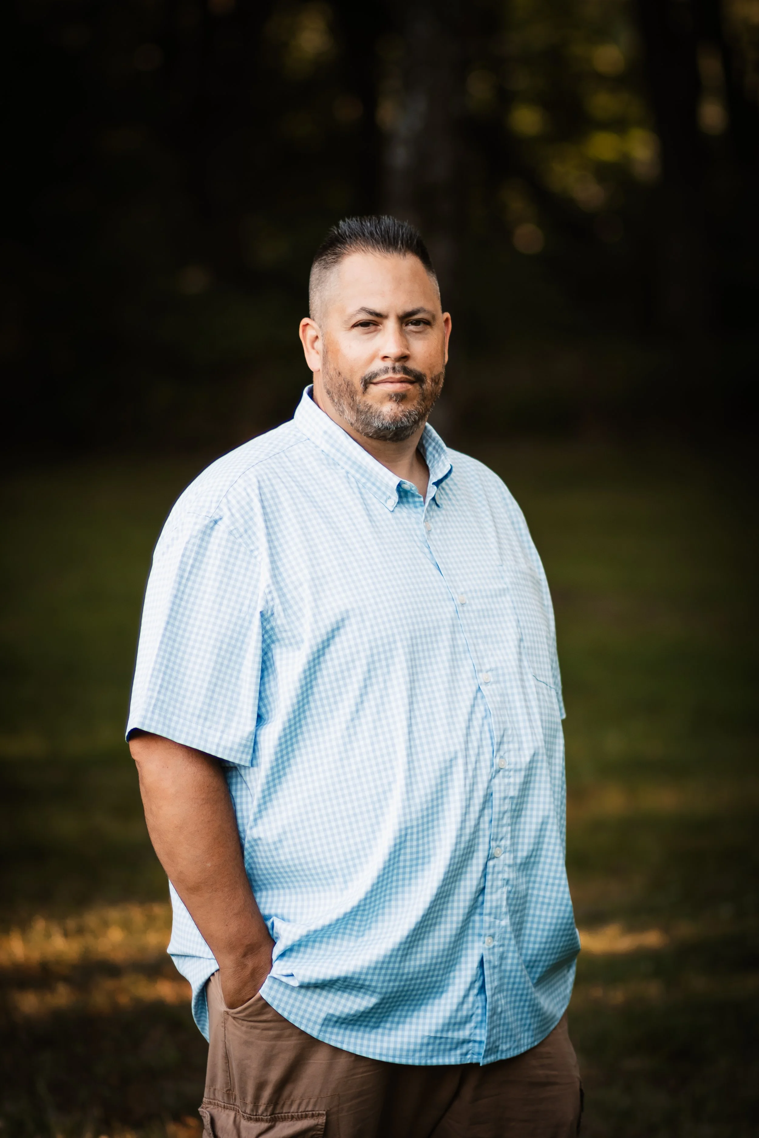 Man with short dark hair and a beard wearing a light blue checked shirt and brown pants standing outdoors in a park at sunset.