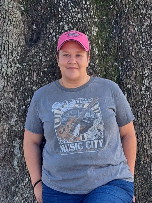 Woman standing outdoors against a large tree trunk, wearing a pink cap and a gray T-shirt with a Nashville music city graphic, smiling.