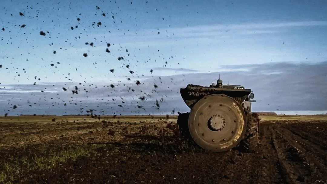 T-Rex Ditcher being pulled by a large tractor with a cloud of dirt and debris being thrown up into the air, under a cloudy sky.