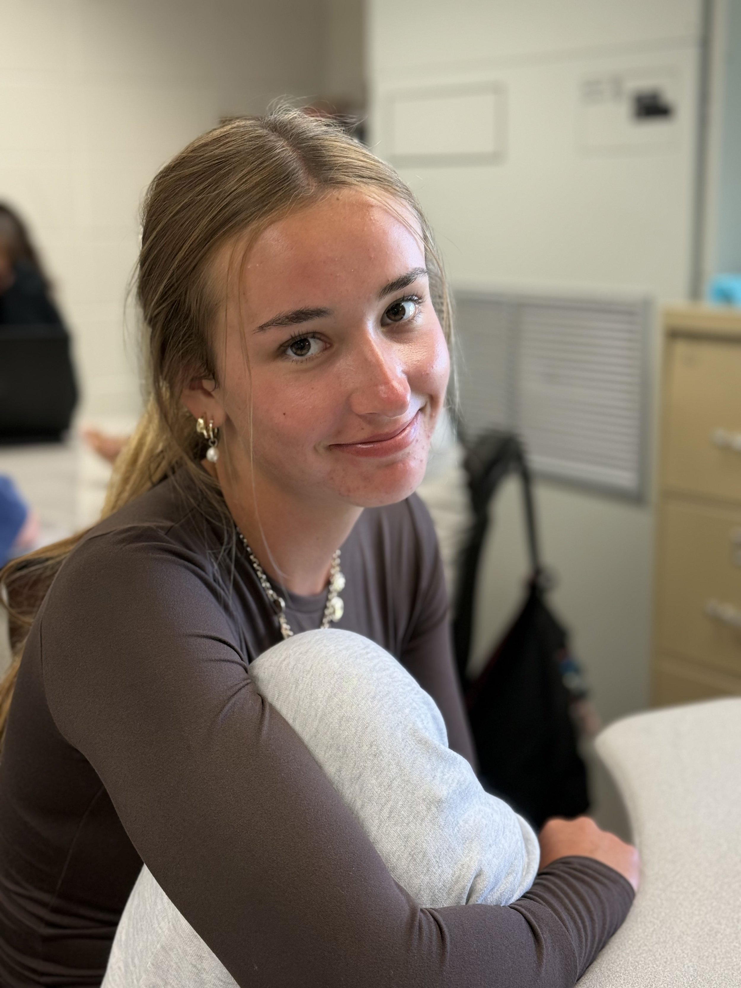 A young woman with long hair, wearing pearl earrings and a necklace, smiling while sitting at a table in an indoor setting.