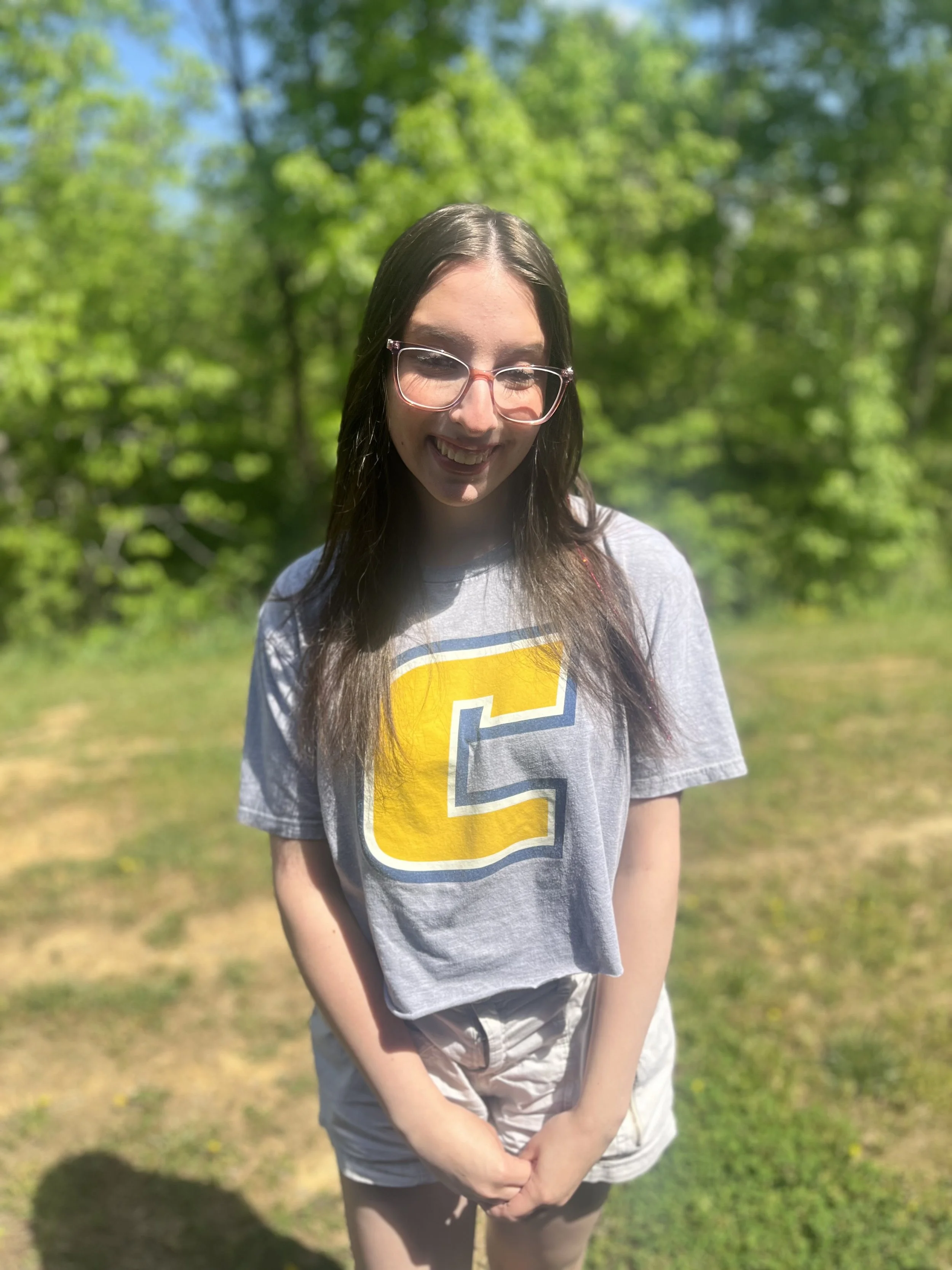 Smiling young woman with glasses and dark hair, sitting at a desk in a classroom, with another person blurred in the background.