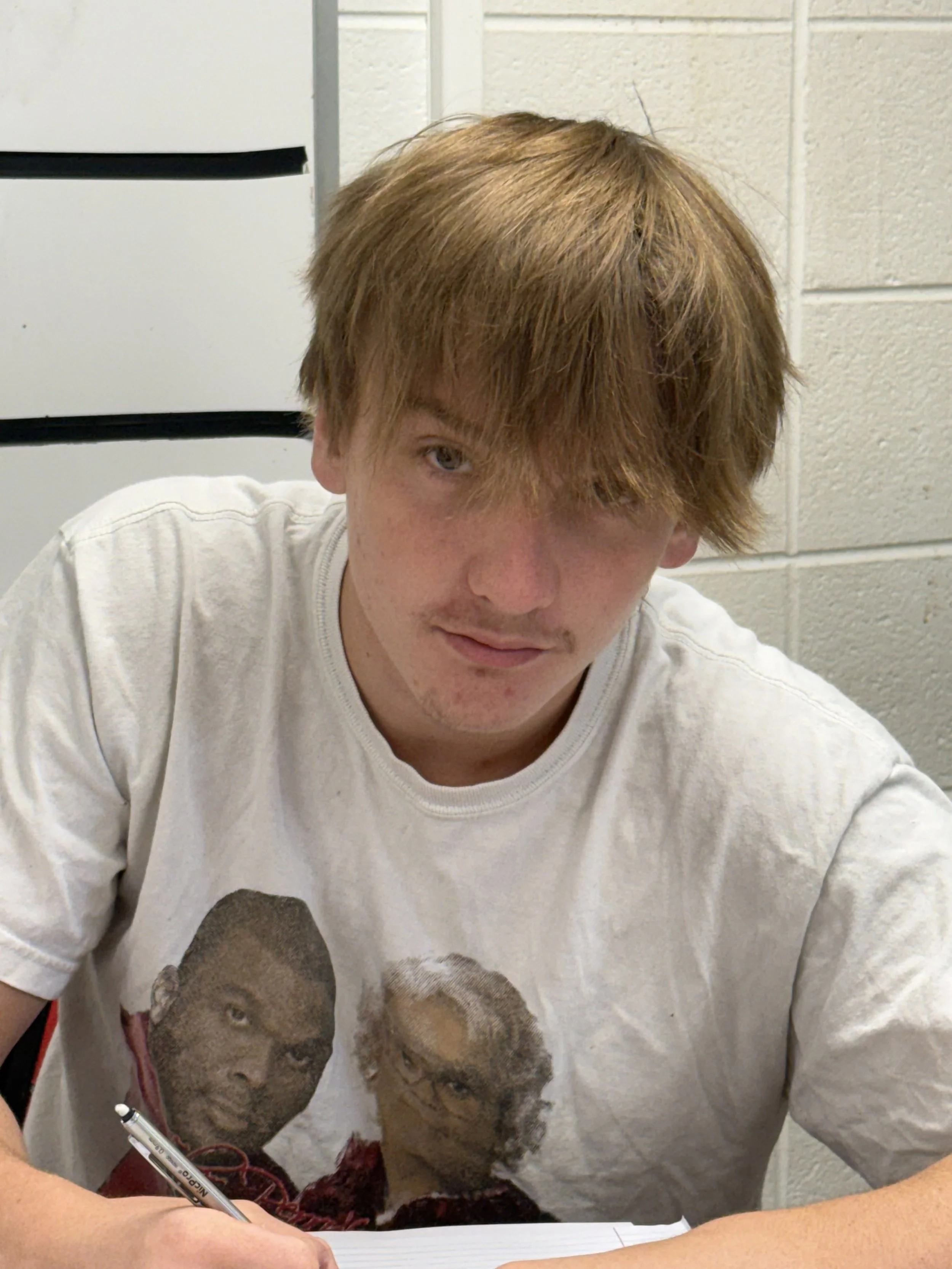 A smiling young boy with short light brown hair, wearing a dark gray T-shirt, sitting in a classroom or office environment with shelves, books, and a desk in the background.