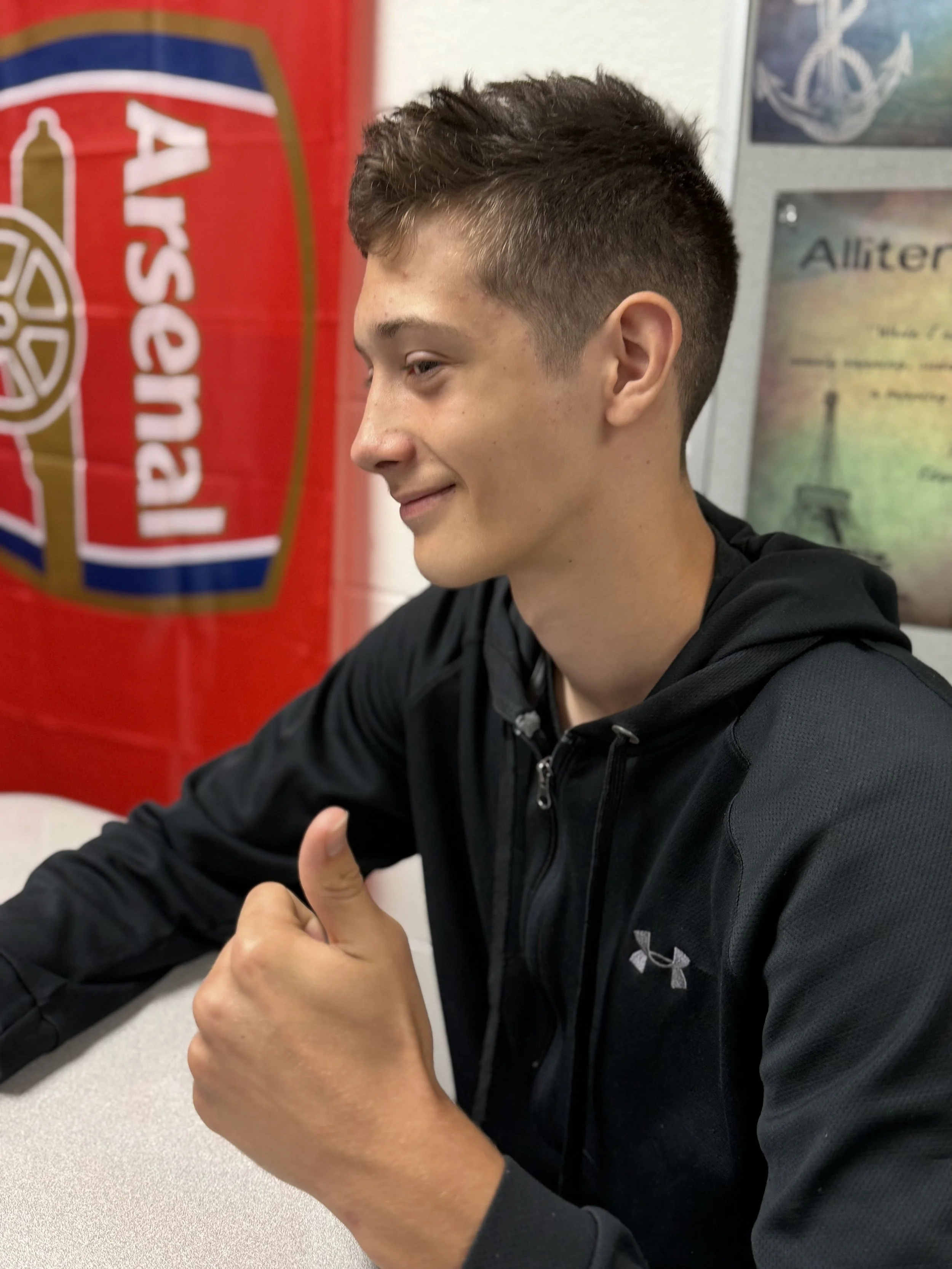 A young man with short brown hair giving a thumbs up while sitting at a table in a room with a red flag and military posters on the wall.
