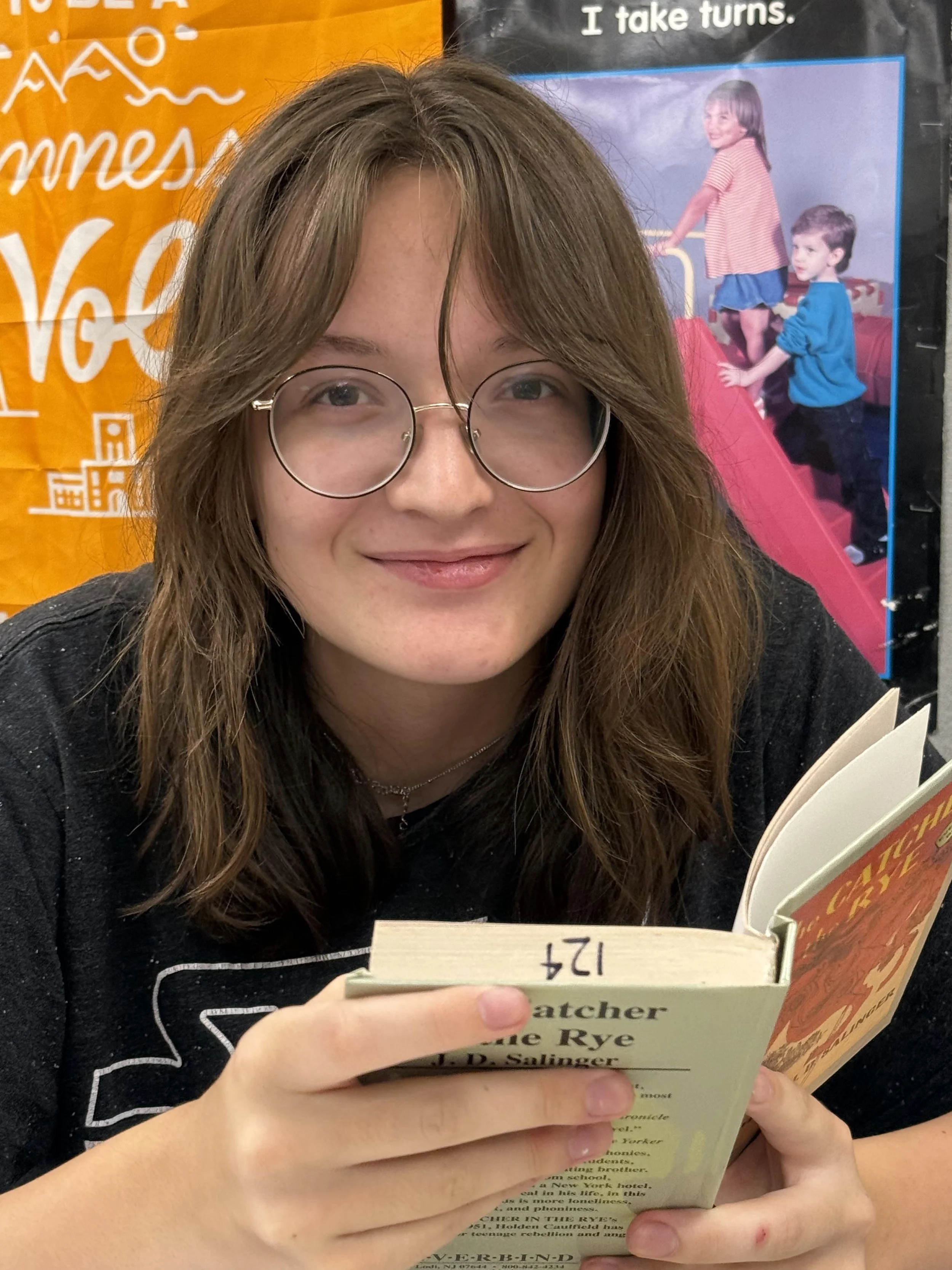 A young woman with brown hair, glasses, and freckles is smiling in a classroom setting.