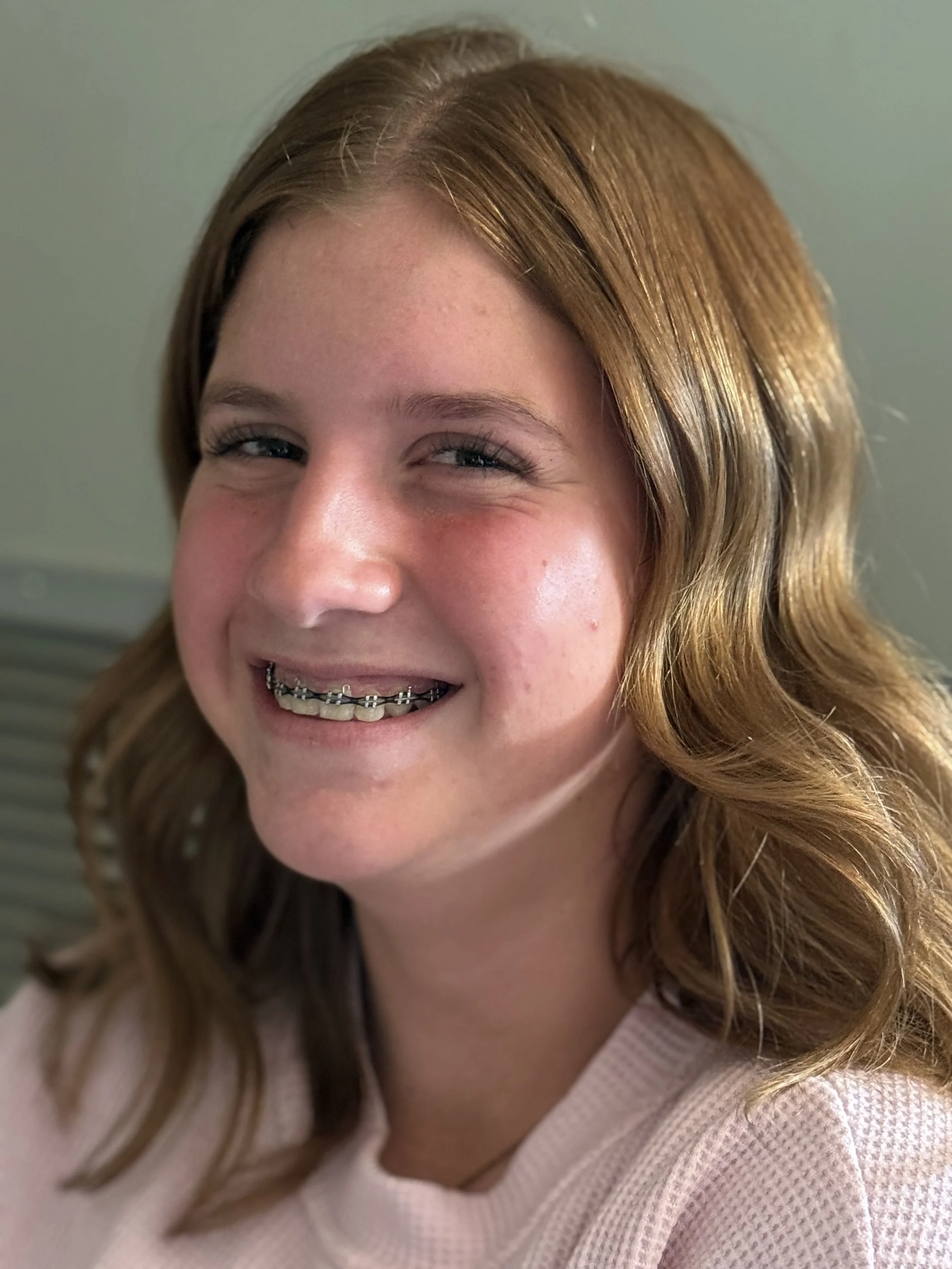 Close-up of a smiling young woman with red hair, braces, and bright eyes, wearing a light pink shirt.