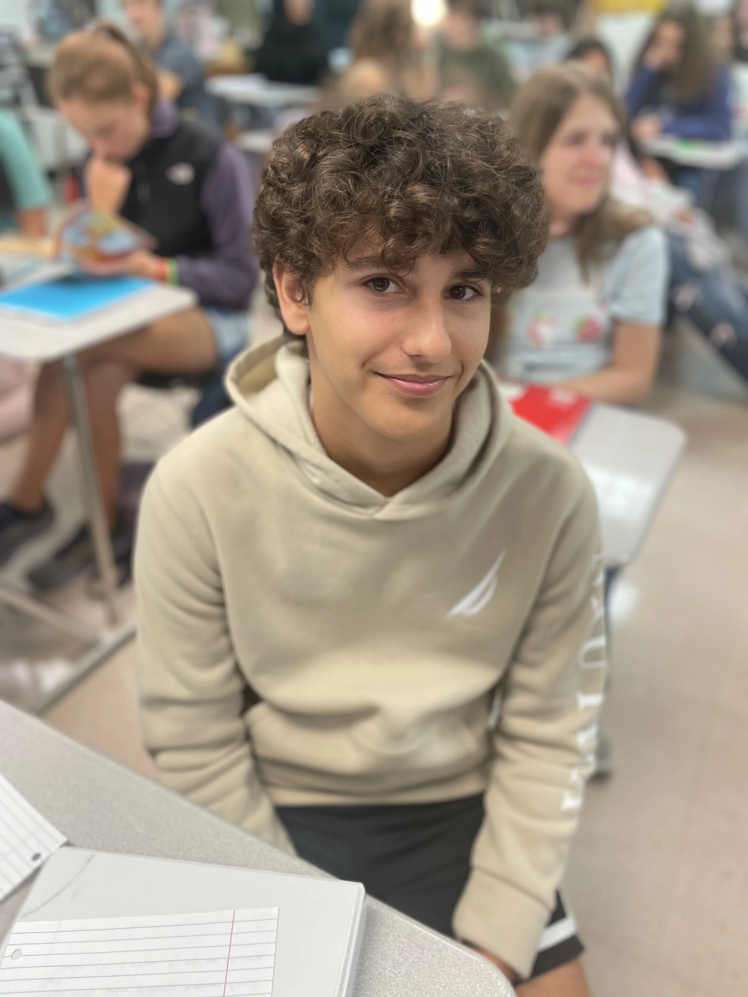 A boy with curly hair and a beige hoodie sitting at a desk in a classroom, with other students in the background.